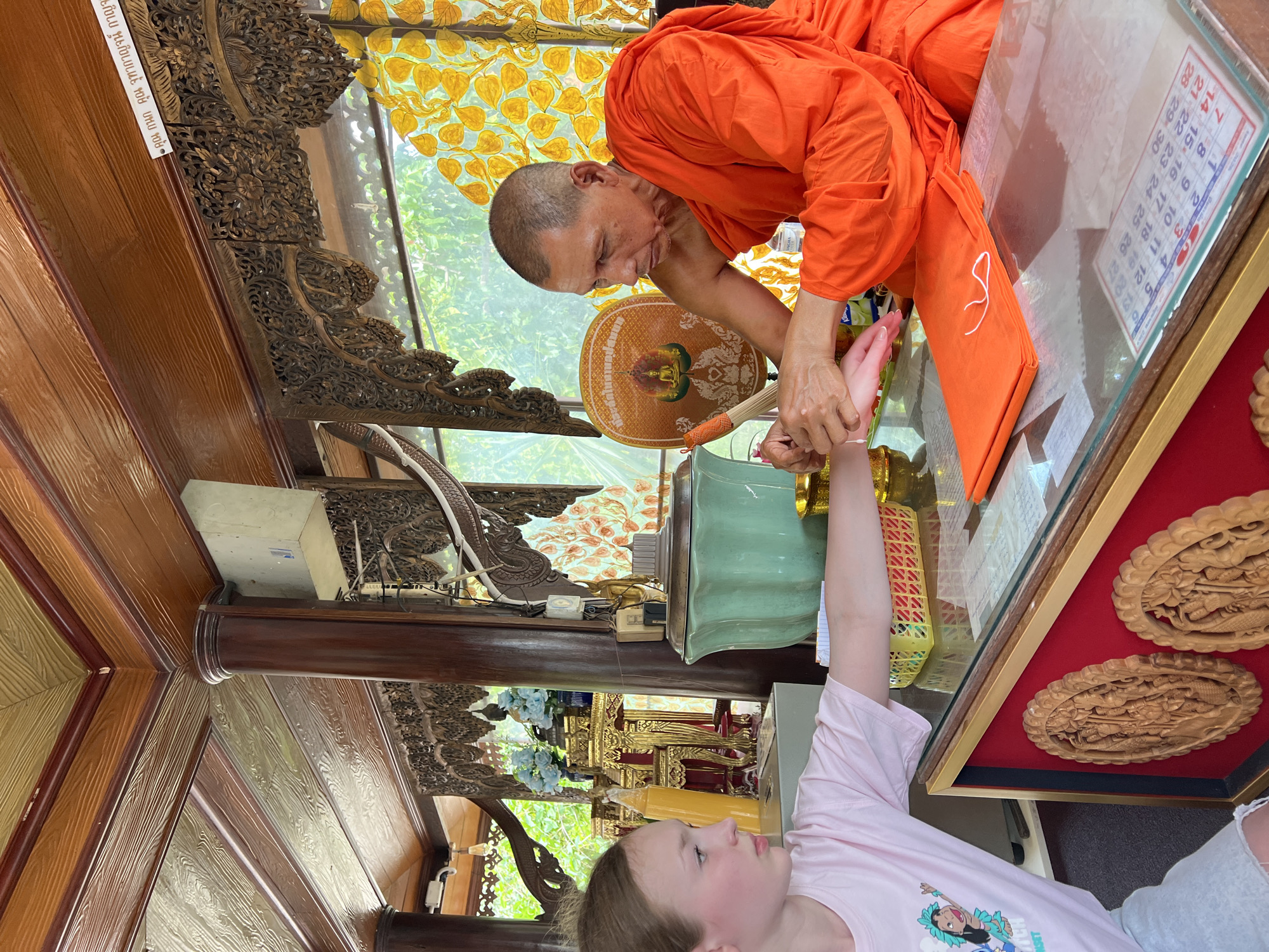 In the city of Ban Bang Rak, Thailand, a Buddhist monk in an orange robe is performing a ritual on a young girl. The monk is holding the girl's wrist and appears to be applying something or tying a string. The girl, wearing a pink t-shirt with a cartoon character, is looking towards the monk. The scene takes place indoors, in what appears to be a temple or shrine area. The background features ornate wooden carvings, religious statues, and a large green urn. A folded orange cloth is visible on the table in front of the monk.