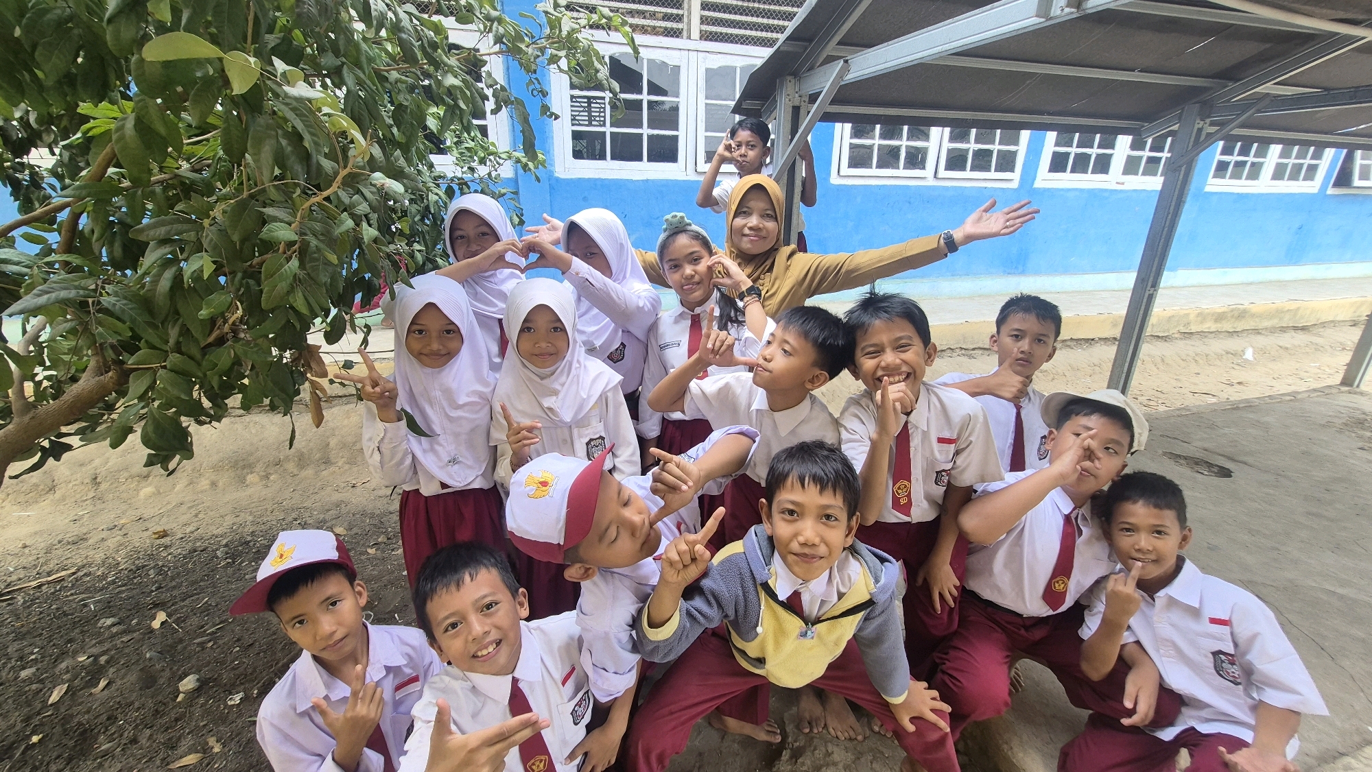 In Putak, Indonesia, a group of schoolchildren, some wearing white uniforms with maroon skirts and ties, gather outdoors in front of a bright blue building. A few girls in hijabs are among the group. One adult woman, wearing a mustard-colored top, is positioned behind the children and appears to be interacting with them. Many of the children are making hand gestures, some forming heart shapes, others pointing or giving peace signs, suggesting a posed, playful moment. A tree with dense green foliage is to the left of the frame. The ground is a mix of dirt and pebbles. A covered shelter with a dark roof and metal poles is visible in the background.