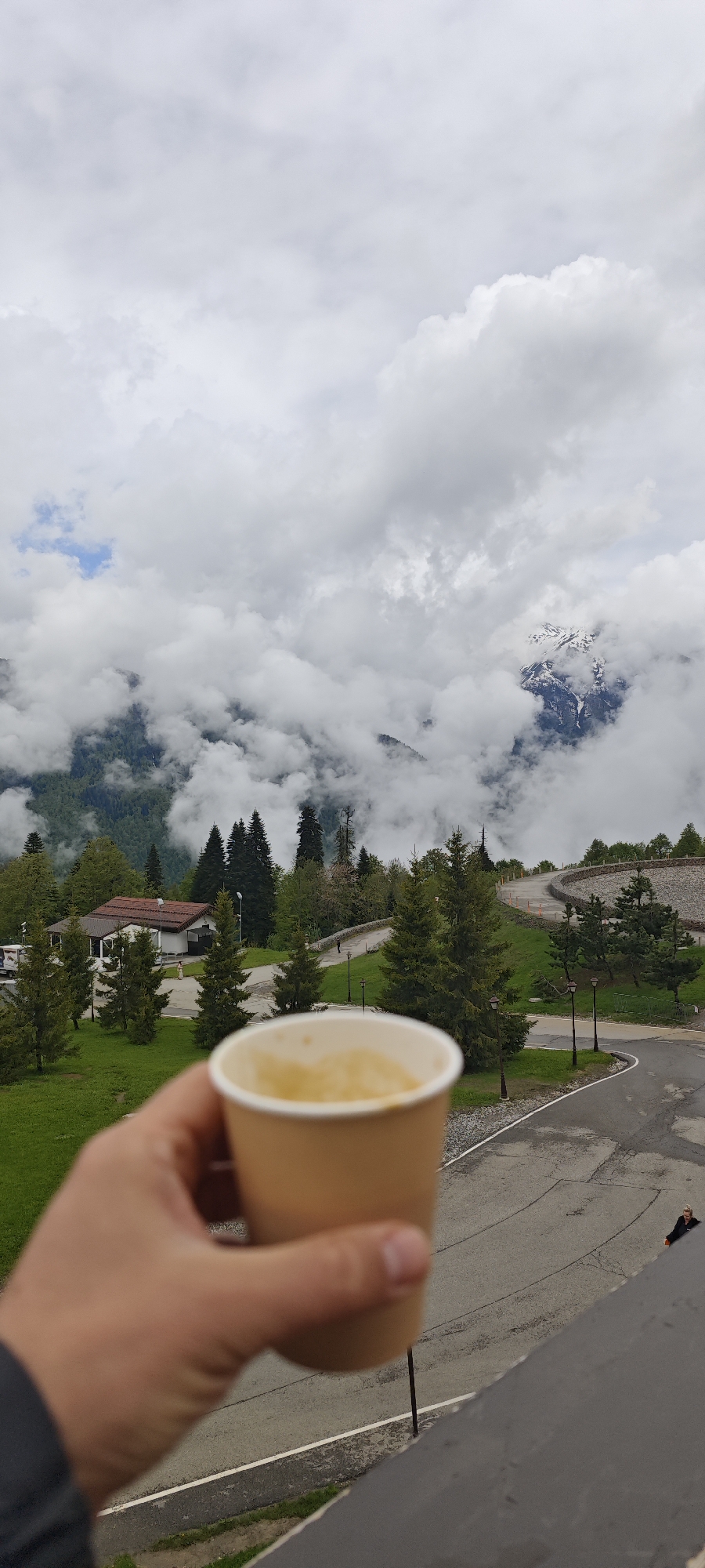 A person holds a paper cup of a yellow-colored liquid in front of a landscape. The location is possibly in Unknown, Russia. The background includes a building, trees, roads, and a mountain range with clouds. A road winds through a green lawn with light poles. Patches of snow are visible on the mountains.