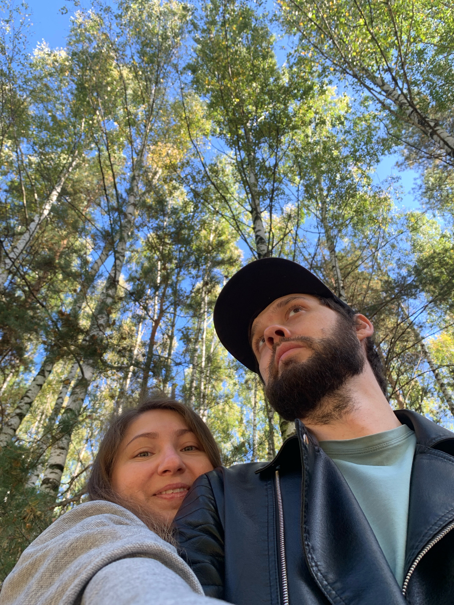 A selfie taken from a low angle shows two people in a wooded area with birch trees against a blue sky. The woman in the foreground, on the left, is wearing a gray knitted sweater and is smiling at the camera. To her right, a man with a beard and wearing a black baseball cap and a black leather jacket over a light blue t-shirt looks up and to the right. The image appears to have been taken in a park or natural setting in Chaikovsky, Russia.