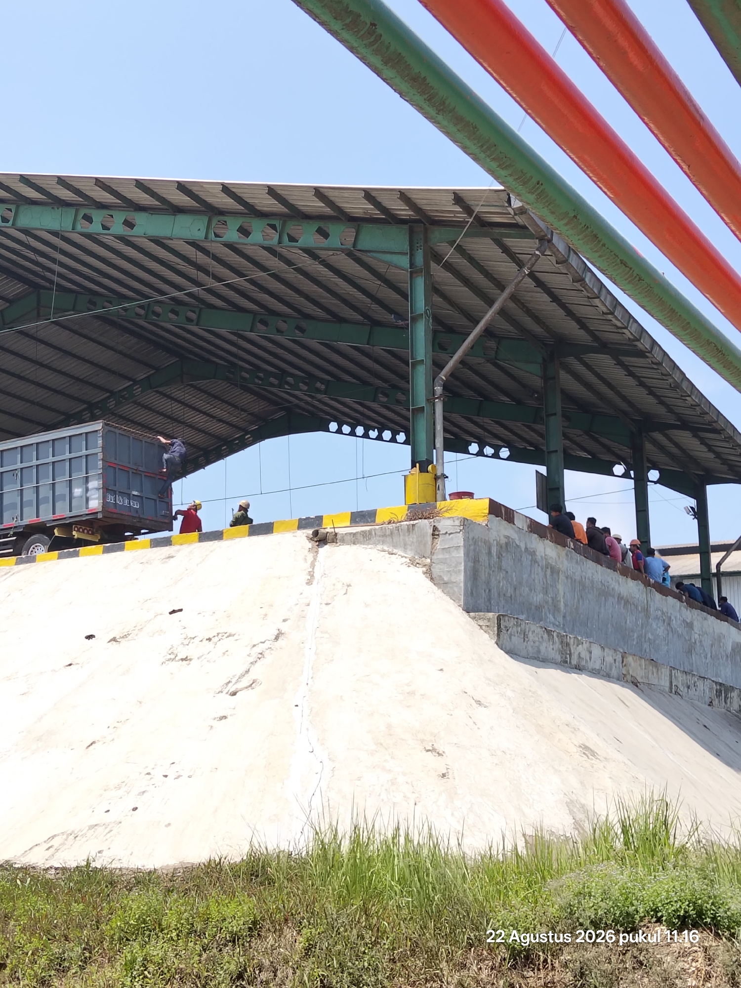 The image depicts an outdoor scene under a clear blue sky, likely a weighbridge or loading area. A large, grey truck trailer is parked beneath a covered structure supported by green metal beams. A person is seen climbing onto the side of the trailer. To the right of the truck, a group of people, some wearing hard hats and high-visibility vests, are gathered on a concrete platform. The platform is bordered by a yellow and black striped safety line and a grey concrete wall. A sloped, light-colored embankment, possibly part of a ramp or foundation, dominates the foreground. Green grass and vegetation grow at the base of the embankment. In the upper right of the image, large, colorful pipes, red and green, are visible. The lower right corner of the image displays a timestamp: "22 Agustus 2026 pukul 11.16".