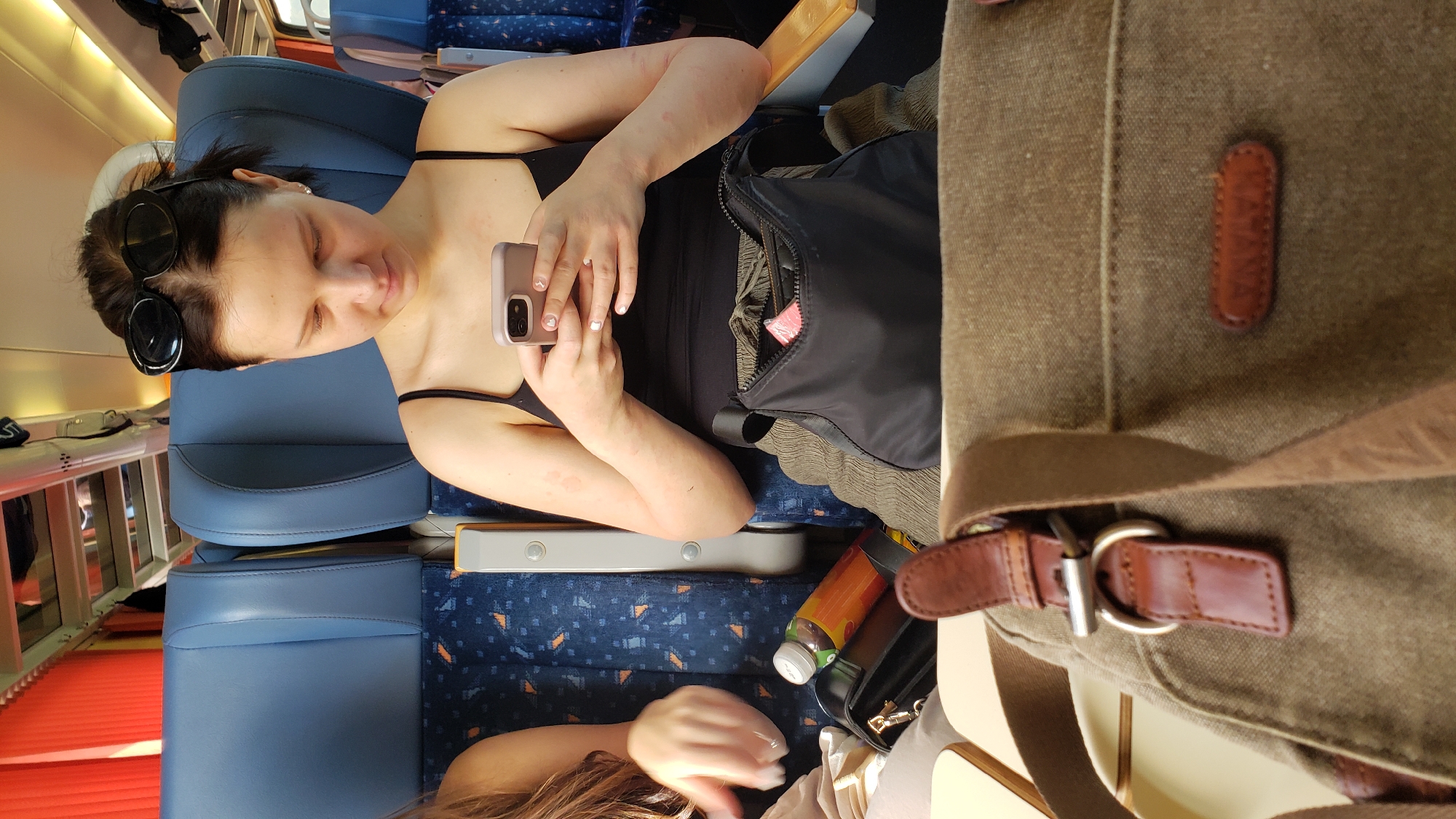 A woman, with dark hair pulled back and sunglasses resting on her head, sits on a train. She is wearing a black tank top and green pants, and is holding a smartphone in her hands, looking down at the screen. Her arms have some red marks on them. A black purse rests on her lap, with a pink item protruding from it. A brown canvas bag with leather accents is in the foreground. Behind her, blue train seats are visible. The scene is lit with artificial light. There is no indication of the specific location in Púchov, Slovakia.