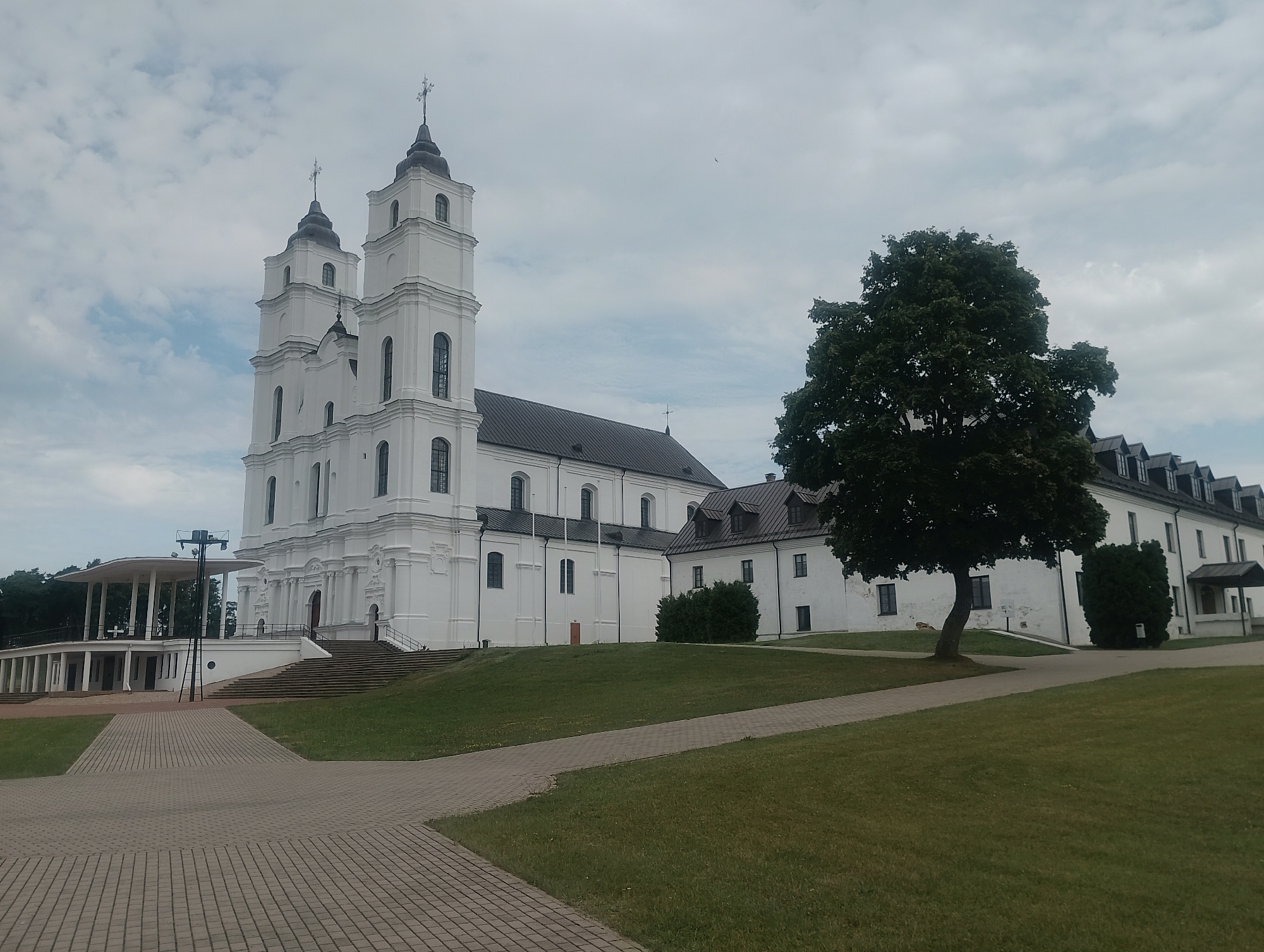 This is a full, eye-level, outdoor, medium shot of the Aglona Basilica and its adjacent buildings, located in Aglona, Latvia. The basilica, a large, white Baroque-style Roman Catholic church, is the dominant subject, featuring two prominent bell towers topped with crosses. The basilica has a dark, sloped roof and numerous arched windows. To the right of the basilica is a long, white building with a dark, mansard-style roof, featuring dormer windows. A large, mature deciduous tree with dark green foliage stands between the basilica and this building. In the foreground, a paved pathway made of grey interlocking bricks curves through a grassy area. To the left of the basilica, a covered, open-air structure with a flat roof supported by columns is visible, with stairs leading up to it. The sky is overcast with scattered clouds. No people are visible in the scene. The overall impression is one of architectural grandeur set within a peaceful, green landscape.