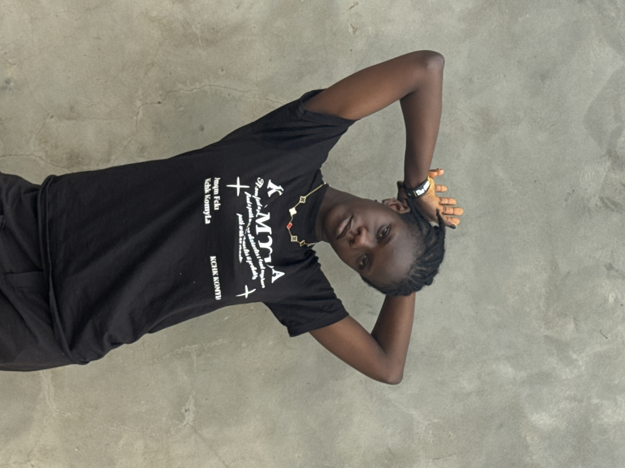 A young person is posing in a photograph. The individual is wearing a black t-shirt with white print and dark pants. They are angled toward the camera and have their hands behind their head. The surface they are standing on appears to be gray and cracked, possibly concrete. The setting is outside, but the specific location in Badagry, Nigeria, cannot be determined from the provided image. There are no other identifiable people, objects, or interactions visible in the image.