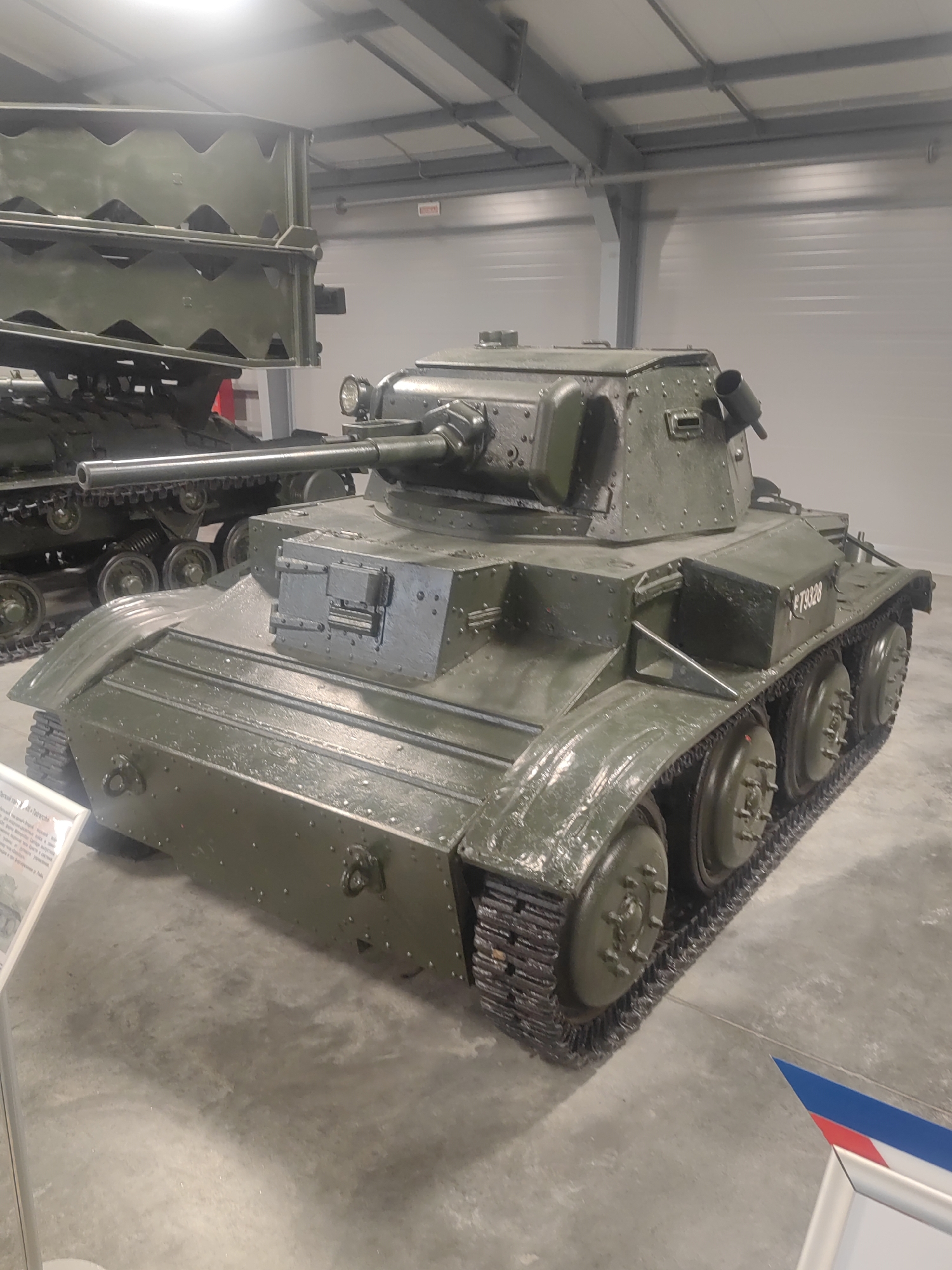 This media file features a military tank, specifically a British Covenanter tank, displayed indoors, likely in a museum. The tank is painted in a dull green, military camouflage. Its tracks are visible, as are its road wheels and turret. A long gun barrel extends from the turret. The tank appears to be in good condition, suggesting it is a preserved exhibit.
In the background, another military vehicle is partially visible, possibly a bridge layer tank, also in a similar green color. The setting is a large, indoor exhibition hall with a high ceiling supported by metal beams. Fluorescent lighting illuminates the scene from above. The floor is concrete, showing some scuff marks and dust.
To the left of the tank, a small display case with text and an image is present, likely providing information about the tank. In the lower right corner, a small section of a tri-colored ribbon, possibly representing the Russian flag, is visible. The overall atmosphere is quiet and educational, typical of a museum exhibit. There is no indication of people or any activity taking place other than the static display of military hardware. The lighting suggests it is daytime, but the indoor setting makes external weather cues irrelevant. One notable detail on the turret of the Covenanter tank is a numerical marking, "F73328". The text in the display case is not legible in the provided image.
