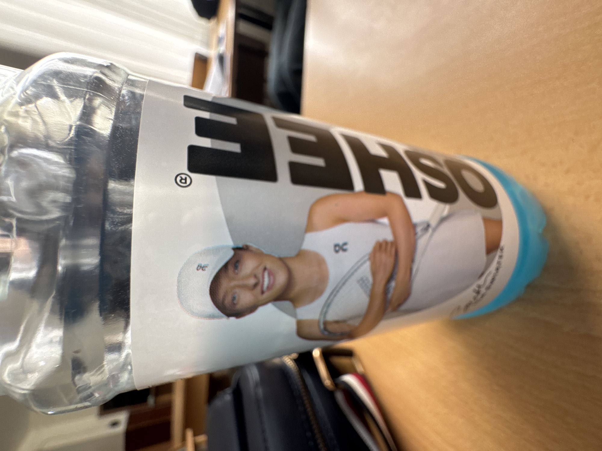 A plastic water bottle, with a label featuring the brand "OSHEE" in large, bold black letters, lies horizontally on a light brown wooden surface. A tennis player, wearing a white tennis outfit and holding a racket, is depicted on the label. The cap of the bottle is blue. A black bag with gold hardware is partially visible in the lower part of the frame. The location in Yerevan, Armenia is not discernible from the image.