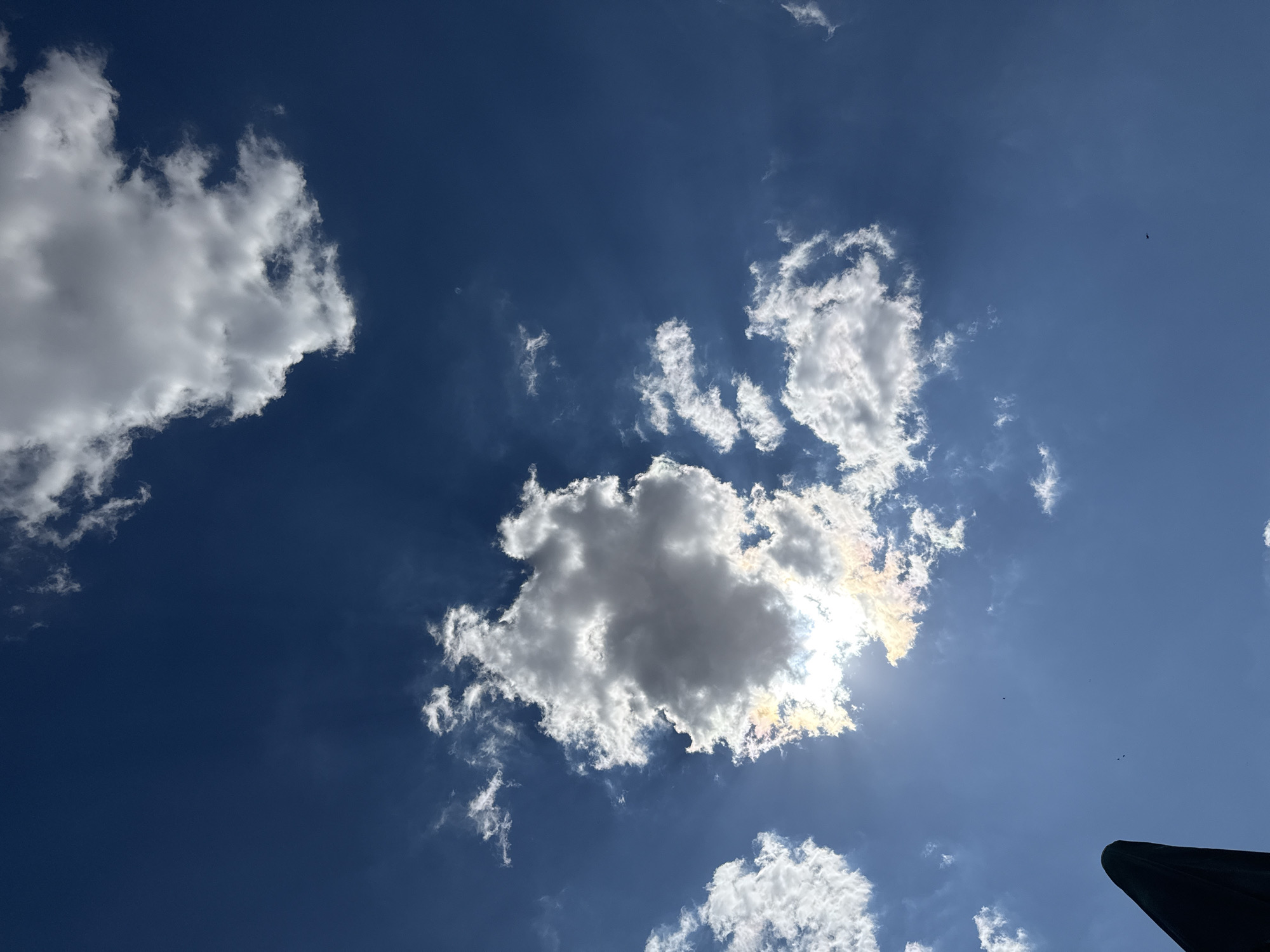 The image shows a deep blue sky with several white clouds. The sun is partially obscured by a large cloud in the center-left of the frame, creating a halo effect with rainbow colors around its edge. A dark, angular object, possibly the top of an umbrella or a structure, is visible in the lower left corner. There are also a few small dark specks in the sky, which could be birds or insects. The scene appears to be outdoors during the daytime, likely in a location with a clear sky. No people are visible. The specific location in Ekzarh Antimovo, Bulgaria, is not discernible from the image itself.