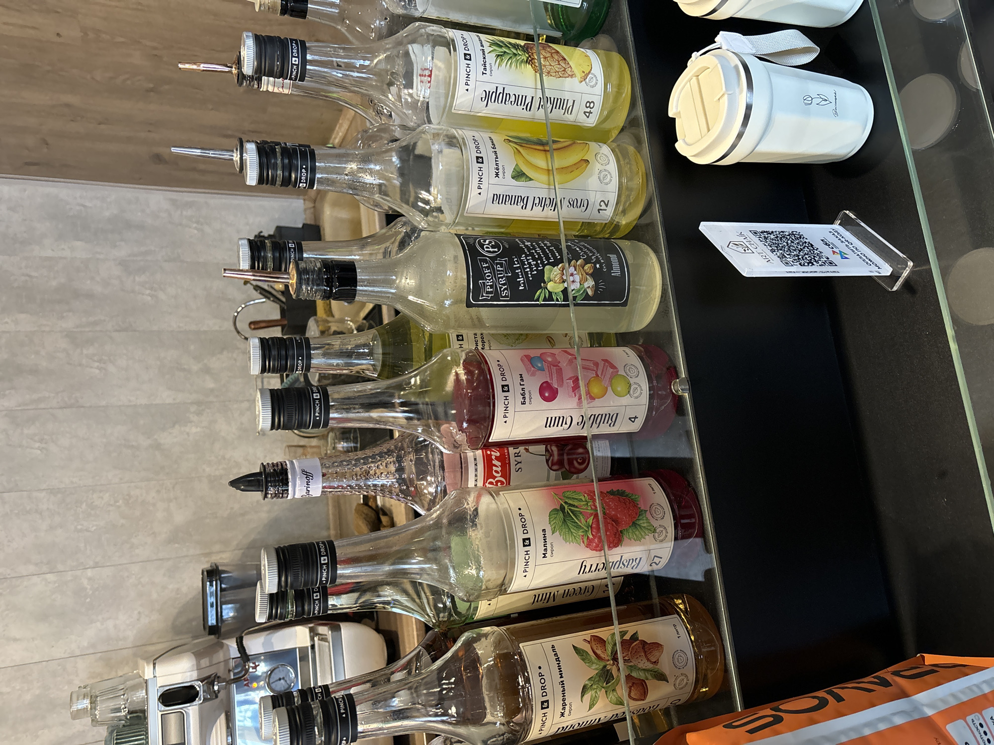 The image displays a collection of syrup bottles arranged on a dark counter, likely within a cafe or beverage-serving establishment in Chaikovsky, Russia.
The main subjects are multiple glass bottles, mostly equipped with metal pour spouts, neatly lined up on a display shelf. Most bottles are from the brand "PINCH & DROP", with some labeled "PROFF SYRUP" and "Barinoff". Visible syrup flavors include "Phuket Pineapple" (Тайский ананас), "Gros Michel Banana" (Жёлтый банан), "Almond" (Жареный миндаль), "Green Mint", "Bubble Gum" (Бабл Гам), and "Raspberry" (Малина). The "PROFF SYRUP" bottle label mentions it's "Perfect for: bright cocktails smoothies lenevades sweet" drinks.
To the right of the bottles, a white insulated mug or tumbler with a grey strap is placed. Next to it, a small acrylic stand holds a card with a QR code and Russian text, which reads "ART CHAIK" and "можно по QR-КОДУ ОПЛАТИТЬ ЗАКАЗ" (you can pay for the order by QR-CODE). In the background on the left, part of a commercial coffee machine or espresso maker with gauges is visible, indicating beverage preparation. A partial orange package with "SON" printed on it is seen in the bottom right. The setting suggests an organized station for customizing drinks with various flavors. The bottles are arranged to be easily accessible, implying an active service area.