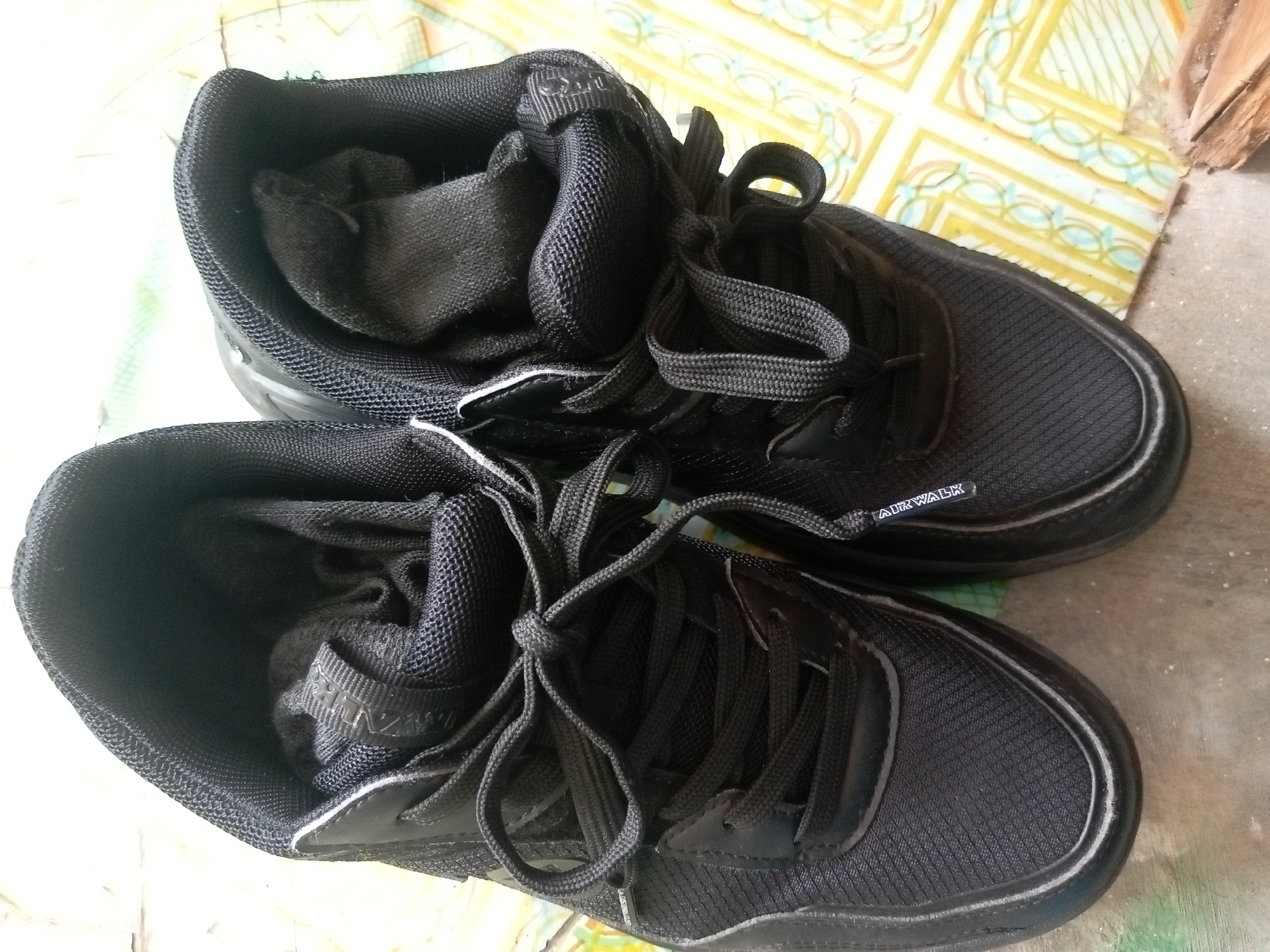 A pair of black Airwalk sneakers with white accents is shown on a tiled floor in what appears to be an outdoor or semi-outdoor setting. The sneakers are placed one on top of the other, with the top sneaker slightly angled. The background features a patterned tile surface and some wood. The image is likely taken in Batam, Indonesia, though specific landmarks are not visible. There are no people present in the image.