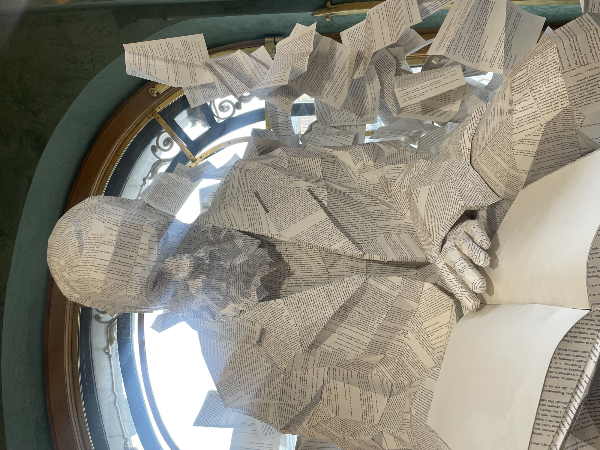 A close-up shot shows a sculpture made of paper, resembling a man with a beard and jacket, sitting in front of a large arched window. The sculpture is covered in text, suggesting it is made from book pages. Loose pages of text are scattered around the sculpture and appear to be flying off from its body and shoulders. The window behind the sculpture has ornate metalwork and a wooden frame. The wall surrounding the window is painted a deep green. The sculpture appears to be an art installation, possibly in a museum or public space. The overall impression is one of knowledge and stories being dispersed.