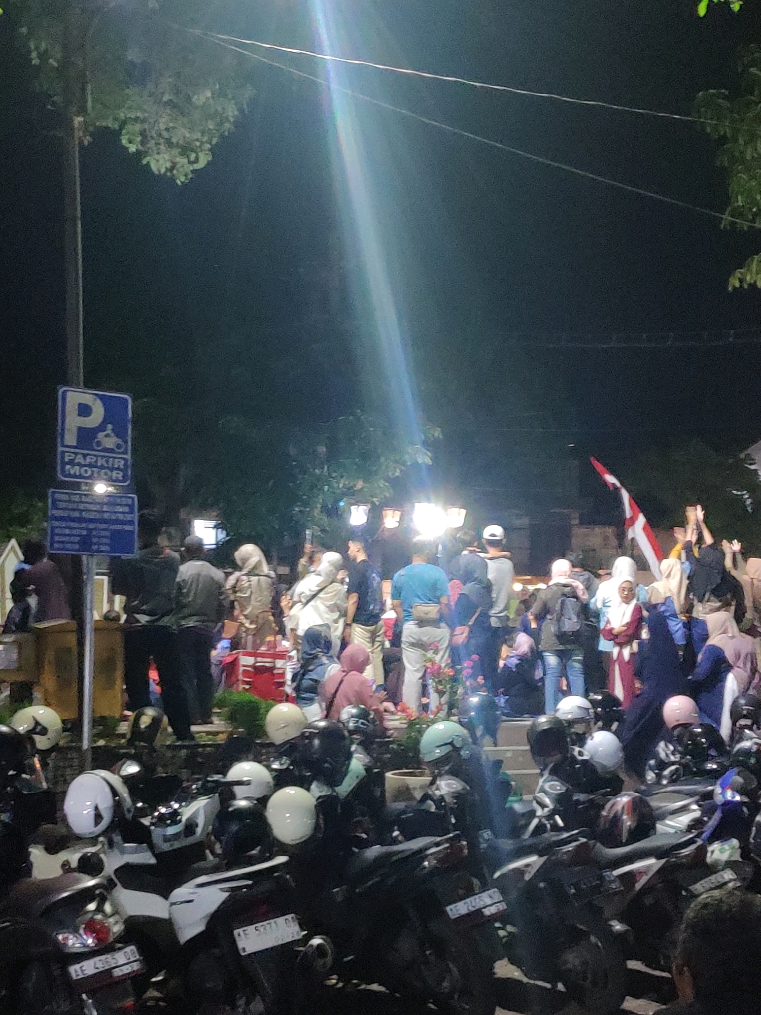 This image captures a nighttime scene in what appears to be a public area in Magetan, Indonesia. A group of people, many wearing headscarves, are gathered, some looking towards the right where a flag with red and white stripes is being held aloft. Several motorcycles are parked in the foreground, with helmets scattered amongst them. A "PARKIR MOTOR" (Motorcycle Parking) sign is visible on the left. Bright lights illuminate the area, casting long shadows and creating a lens flare effect. The environment suggests an outdoor gathering, possibly a celebration or event.