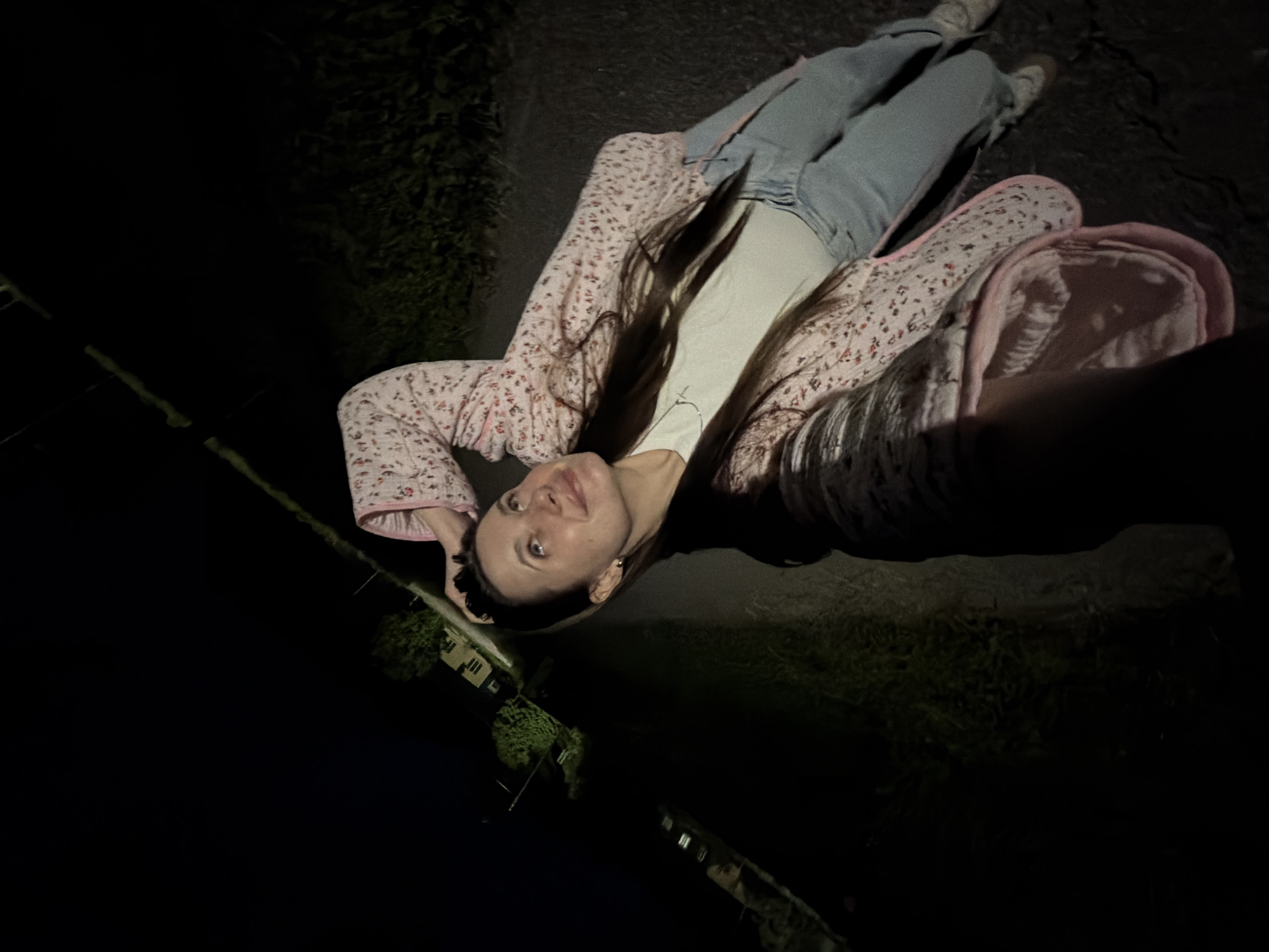 A young woman is standing outdoors at night, possibly in the city of Krasnyy Mayak, Russia. She is looking up and to her left, with her left arm bent and resting on her head. She is wearing a light-colored t-shirt, blue jeans, a pink floral jacket, and white sneakers. The background is dark, with some faint lights from buildings visible in the distance to the left, suggesting a semi-urban or suburban environment. The ground appears to be a paved surface, possibly a road or path. The lighting suggests artificial light sources are illuminating the scene from the front and side.