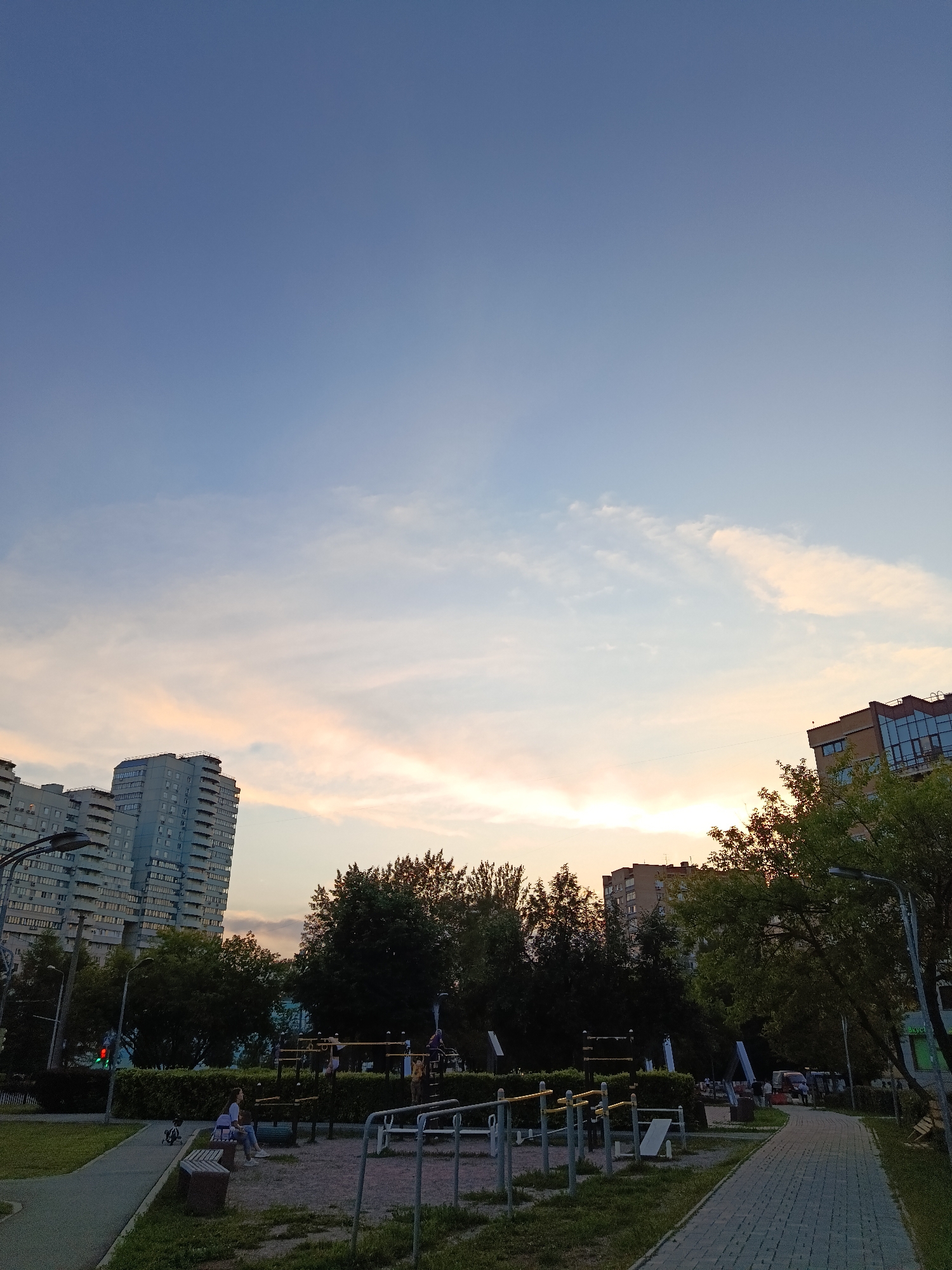 A wide-angle, low-angle shot captures an urban scene at dusk. The sky is a gradient of pale blue to soft pink and orange, with wispy clouds. In the background, apartment buildings of various heights dominate the skyline, some with visible balconies and windows. Trees with dark green foliage are scattered throughout the middle ground, partially obscuring the buildings. In the foreground, a children's playground with metal bars and slides is visible. A paved walkway runs along the right side of the image, bordered by grass. Two individuals are seated on a bench near the playground, and a small child is playing with a toy in the grass. A traffic light with a red signal is also visible in the left portion of the image.