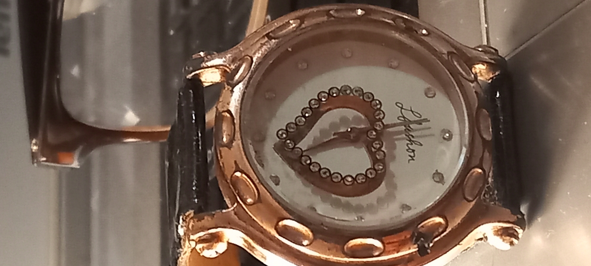 The image displays a close-up of a women's wristwatch. The watch features a rose gold-toned case that shows signs of wear and tarnishing, particularly on its edges and lugs. It has a white or light-colored circular dial adorned with small, clear gemstones or crystals marking the hour positions, although not all hours are marked this way. A prominent design element on the dial is a large heart shape, also outlined with small, clear gemstones/crystals, positioned in the center. The watch hands, appearing in a dark color, indicate the time to be approximately 10:10.
The brand name "J. Leschon" is visible on the right side of the watch face, written in a cursive script. The watch is fitted with a black strap, which also appears to have some wear. The background is out of focus, suggesting the watch is resting on a surface indoors, with hints of other objects or parts of a structure in the blurred areas. There is no specific activity depicted, as it is a still life photograph of the accessory.