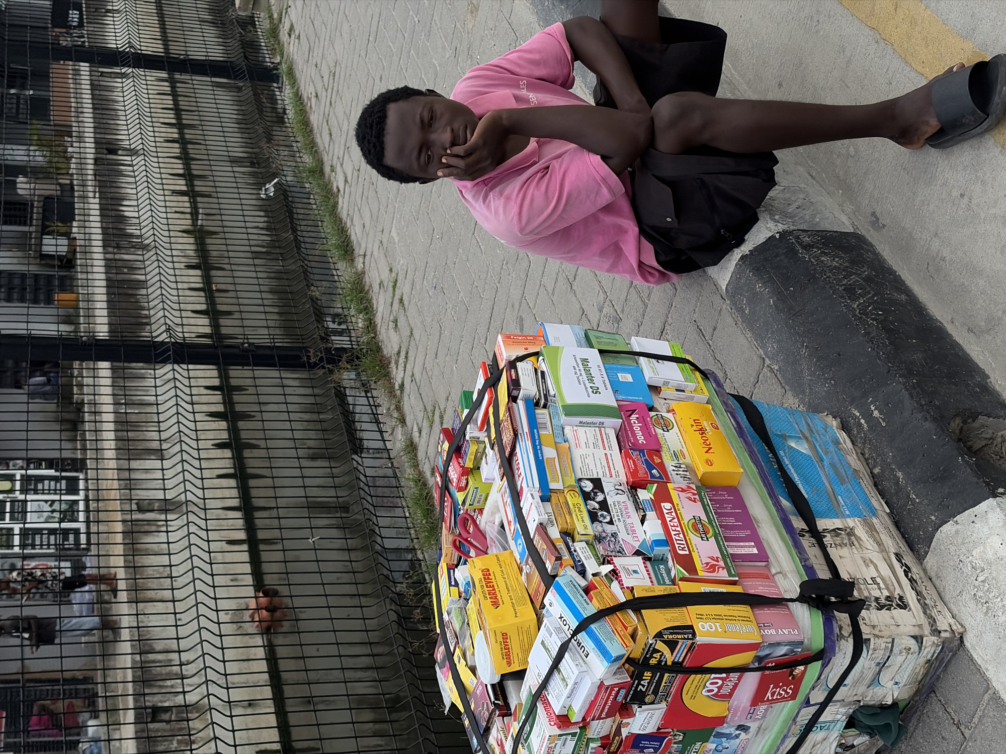 In Igboefon, Nigeria, a young man in a pink polo shirt and black shorts sits on a concrete curb next to a large, stacked collection of packaged medicines. The man rests his chin on his hand, looking down. The stacked medicines, secured with black straps, are on a multi-layered platform, likely a mobile cart. Behind the man and the medicines is a black metal fence separating the sidewalk from a concrete channel containing murky water. Further in the background, through the fence, several individuals are visible standing near a building with multiple windows. The environment appears to be an outdoor urban setting.