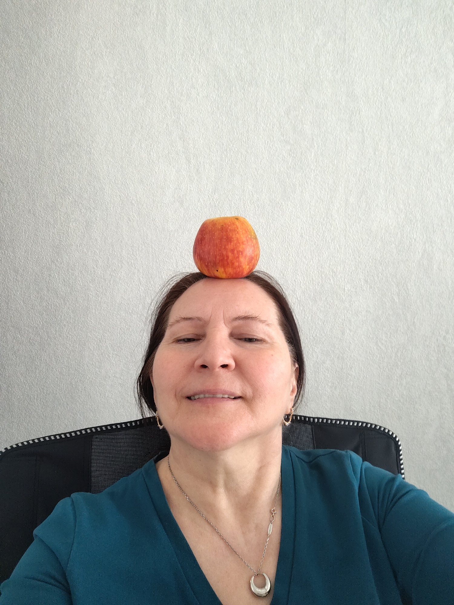 A middle-aged woman is sitting in an office chair against a light gray wall. An apple is balanced on her head. She is wearing a teal top and a silver necklace with a horseshoe-shaped pendant. She has short dark hair and is smiling slightly. The location of this image is unknown, and there is no information suggesting it was taken in Sarapul, Russia.