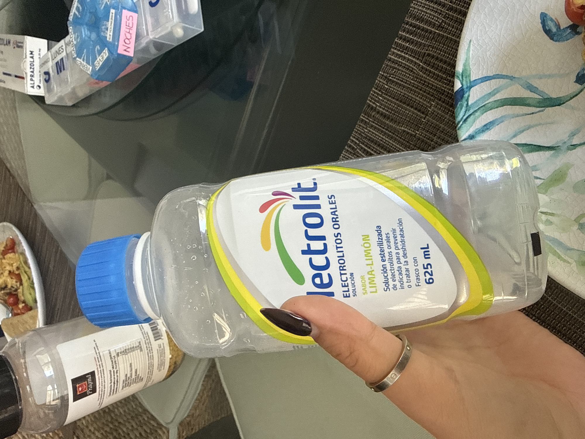 The image captures a close-up, slightly tilted view of a person's hand holding a clear plastic bottle of Electrolit oral rehydration solution. The hand, adorned with dark painted fingernails and a silver ring, holds the bottle prominently. The Electrolit label, featuring a colorful logo, clearly displays "ELECTROLITOS ORALES" (Oral Electrolytes) with "SABOR LIMA-LIMÓN" (Lime-Lemon flavor) and "625 mL". Spanish text on the label specifies it is a "Solución esterilizada de electrolitos orales indicada para prevenir o tratar la deshidratación" (Sterilized oral electrolyte solution indicated to prevent or treat dehydration).
In the immediate foreground to the left, a smaller clear plastic bottle or shaker with a "Pragna" label is visible. The background reveals an indoor setting with various objects on a table. To the far left, a white plate holds a portion of food, possibly a salad with tomatoes. Further into the background, a clear plastic container is labeled "ALPRAZOLAM", and next to it, a blue hexagonal pill organizer shows compartments marked "LUNES" (Monday), "MARTES" (Tuesday), and "NOCHES" (Nights). A patterned ceramic plate with blue and green designs is partially visible on the right. The table surface beneath is dark and appears to have a woven texture. The scene suggests a personal, possibly health-related, context within a home in San Bernardino Tlaxcalancingo, Mexico.
