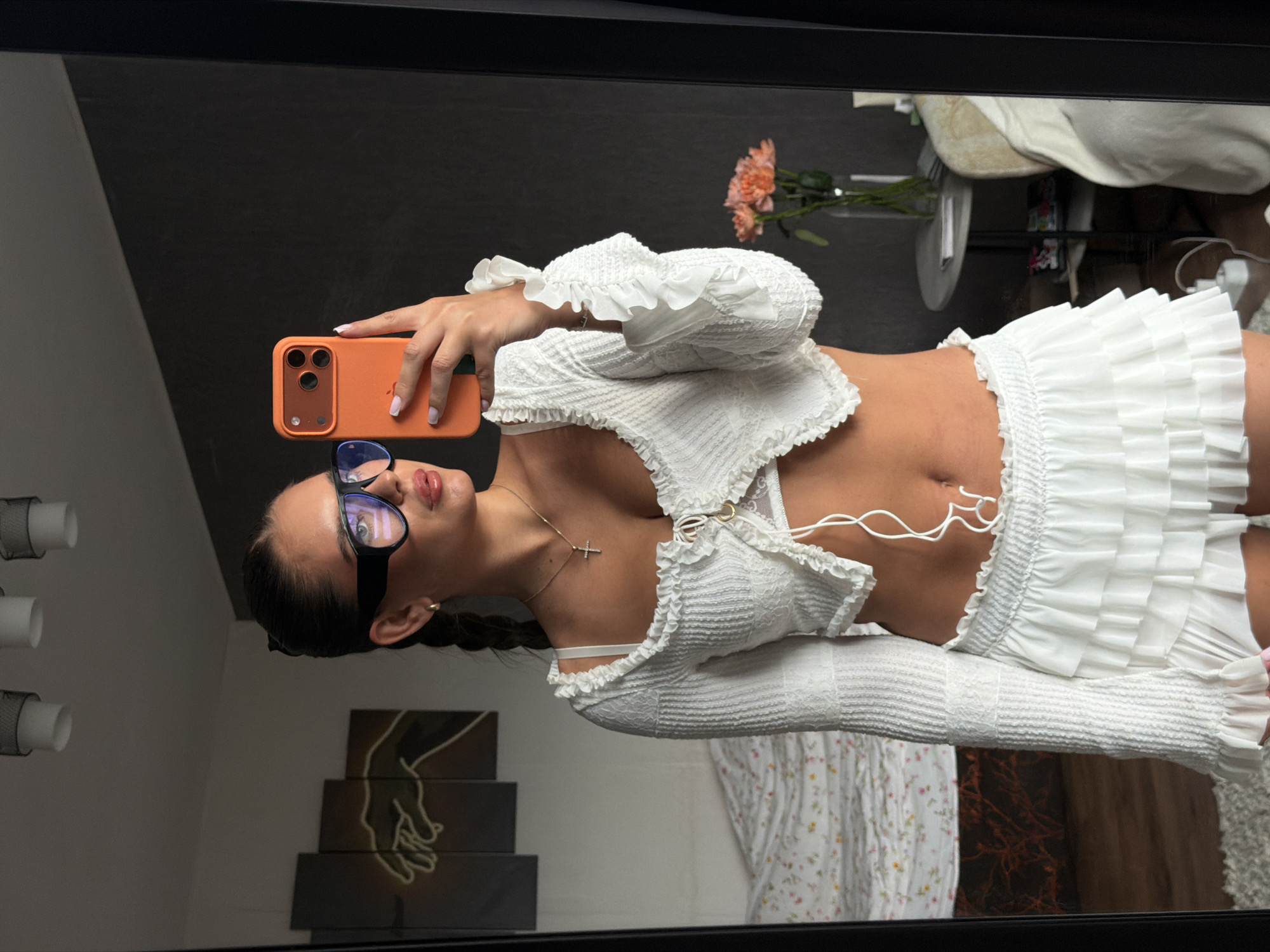 A young woman wearing glasses, a white ruffle-trimmed top and skirt takes a selfie in front of a mirror, holding an orange phone. The background includes interior design elements, such as a lamp, artwork, and a floral arrangement. There is no indication of the city of Krasnoyarsk, Russia.