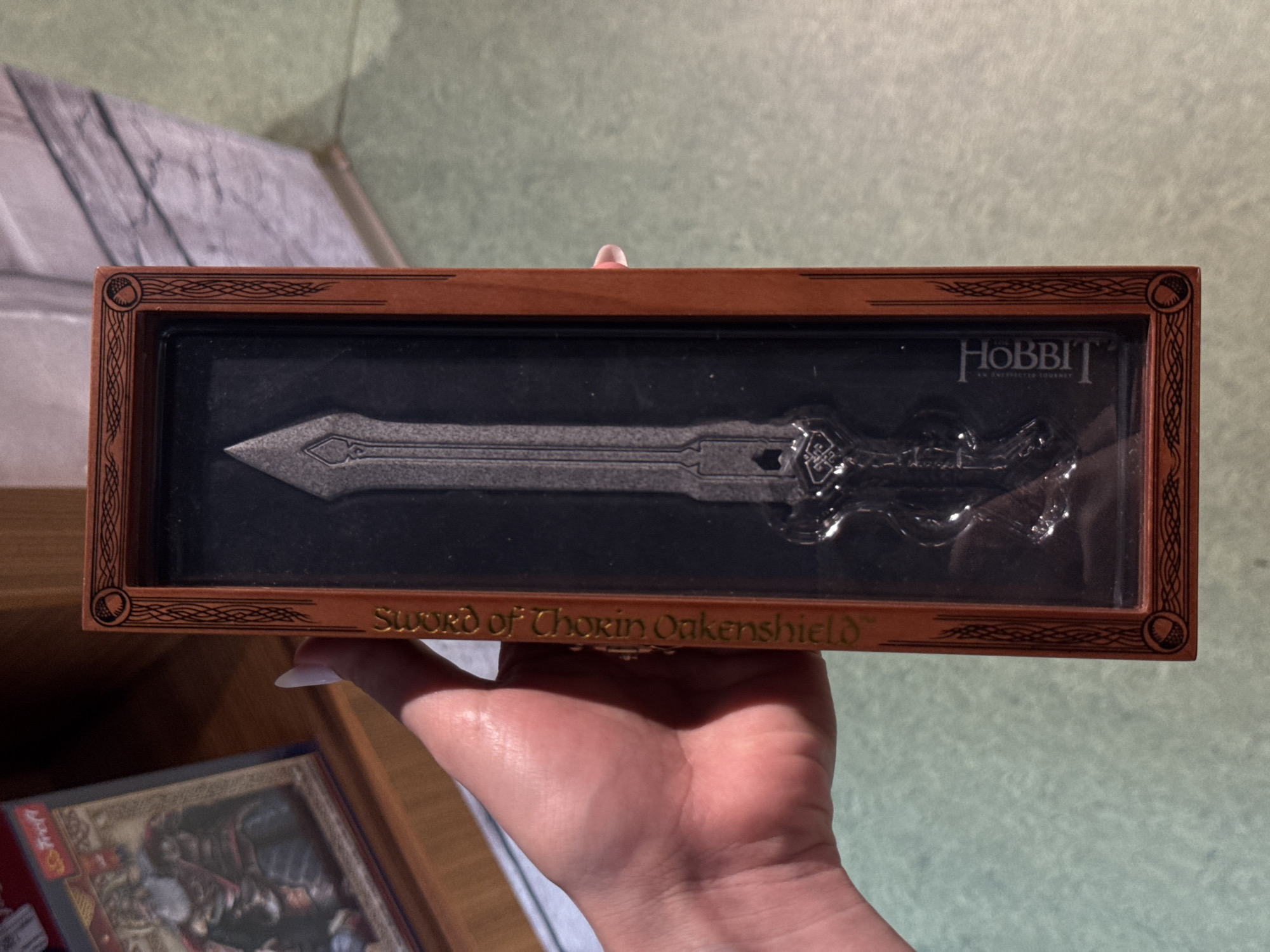 In Yekaterinburg, Russia, a hand holds a framed collectible sword. The sword, labeled "Sword of Thorin Oakenshield" and "The Hobbit," is displayed in a dark, protective case with ornate wooden framing. The background suggests a retail environment, possibly a store shelf displaying other merchandise, with a hint of green carpeting. The hand, with manicured fingernails, is positioned to the left, partially obscuring the left edge of the frame. The lighting is bright, highlighting the details of the sword and the frame.