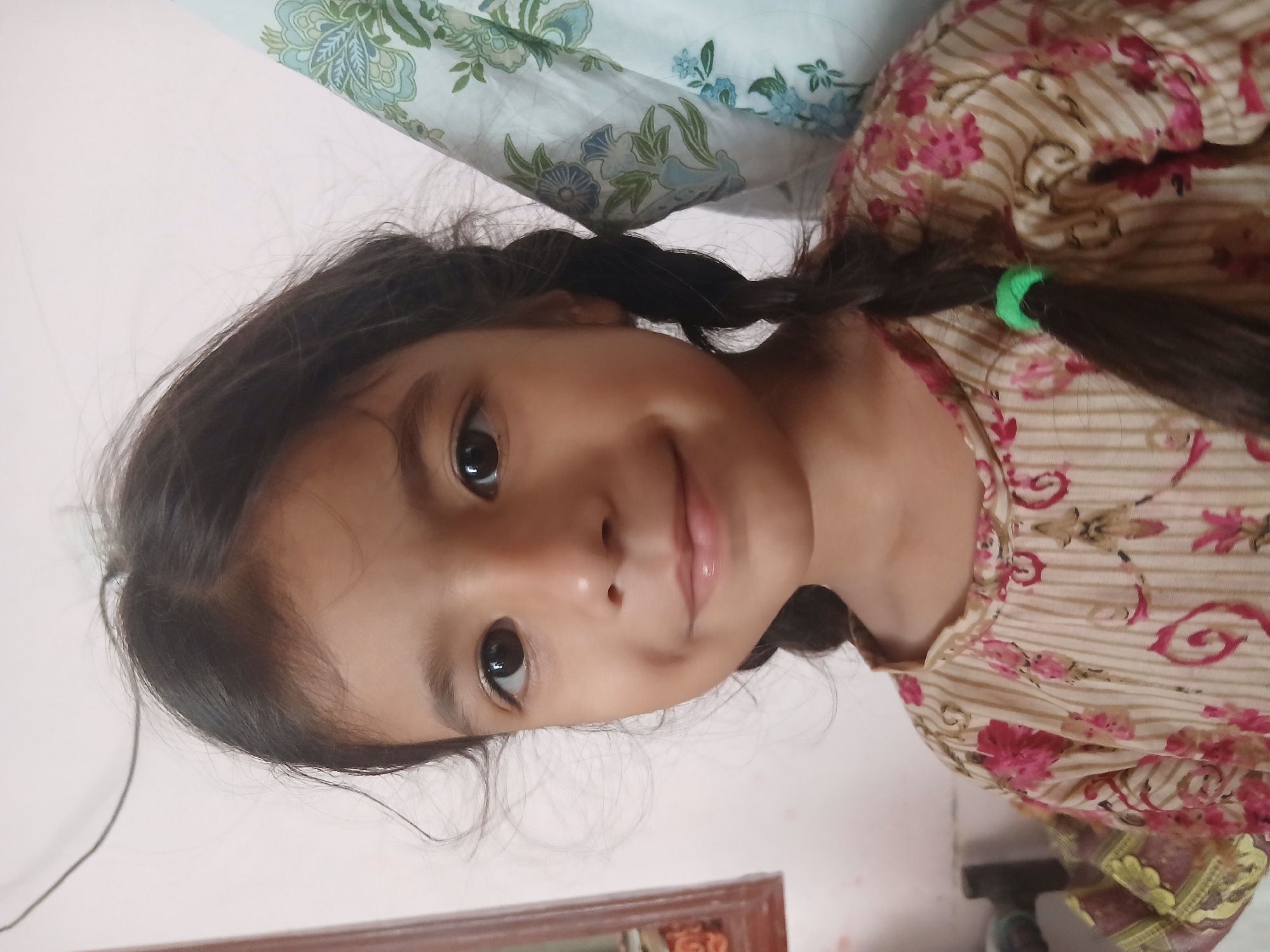 A close-up, head-and-shoulders portrait shows a young South Asian girl with dark hair in two braids. She has dark eyes, a small nose, and a slight smile, with dimples visible on her cheeks. She is wearing a cream-colored shirt with a pattern of vertical brown stripes and scattered pink and red floral designs. Her hair is partially obscuring a lightly patterned fabric to her right, which appears to be a curtain or draped cloth with blue and green floral motifs. To her left, a blurred wooden frame with decorative elements is visible. The background is a plain, light-colored wall, with a dark wire extending diagonally from the upper left corner. The image is taken in Peshawar City Tehsil, Pakistan.