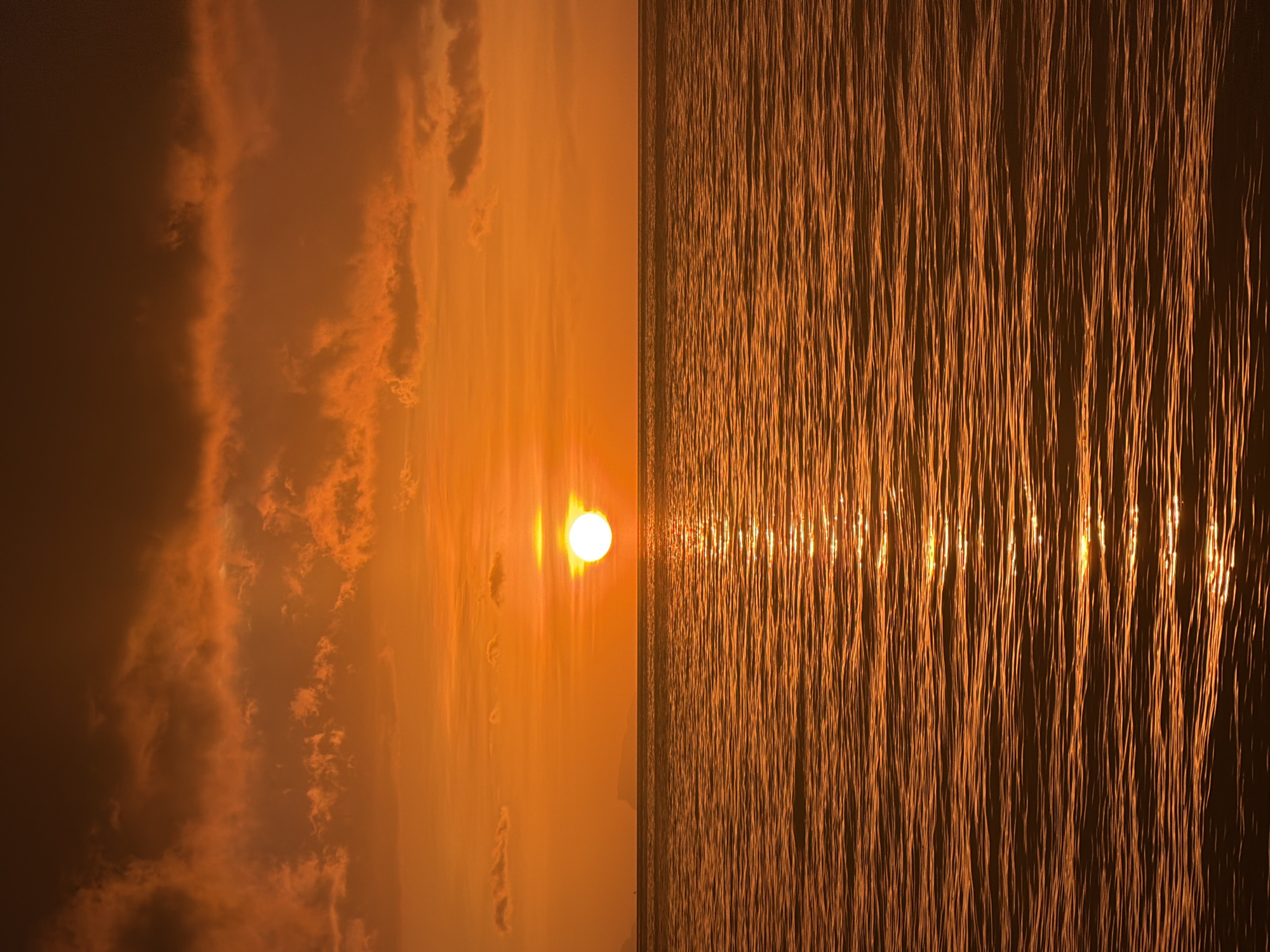 The image depicts a serene sunset over the ocean, casting a warm, orange glow across the scene. The sun, a bright, circular orb, is low on the horizon, partially obscured by wispy, orange-tinted clouds. The surface of the water is gently rippled, reflecting the golden light of the setting sun in a shimmering path. In the distance, the silhouette of land can be seen, hinting at a coastline. The overall atmosphere is tranquil and peaceful. There are no people, animals, or man-made structures visible in the image, suggesting a natural and undisturbed environment. The scene is likely located on the coast of Baan Tai, Thailand, given the typical sunset views and coastal landscape of the area.