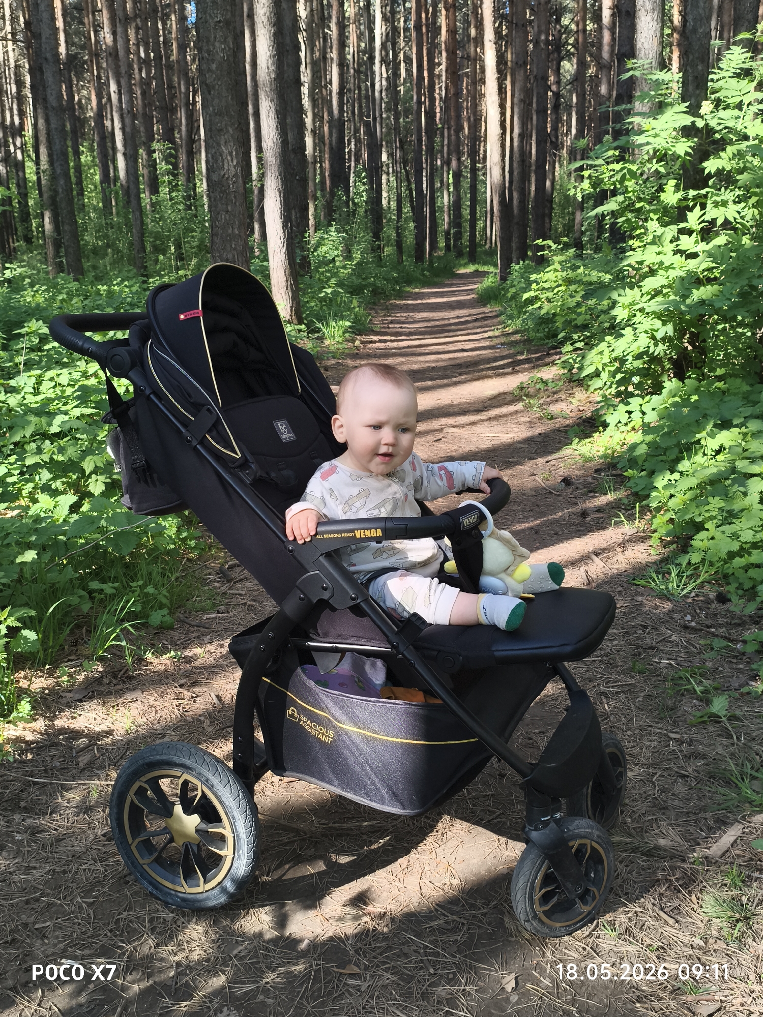 A baby sits in a black and gold Venga brand stroller on a dirt path in a forest. The baby is wearing a light-colored pajama set with a car pattern and is holding a stuffed animal. Tall trees line the path, with green foliage along the sides. The date and time, "18.05.2026 09:11," and the phone model, "POCO X7," are visible in the bottom corners of the image. The location is in the city of Chaikovsky, Russia.