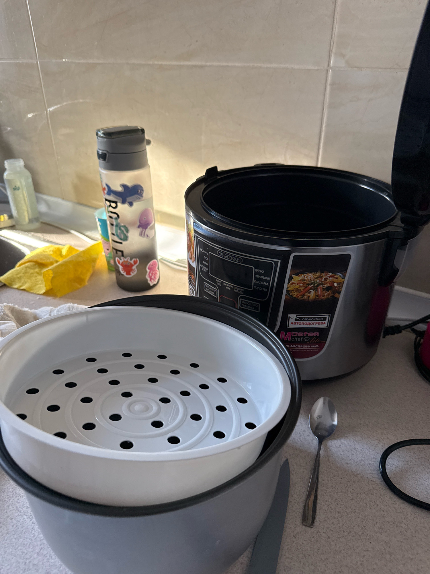The image depicts a kitchen counter with a multicooker, a steaming basket, a water bottle adorned with stickers, and a spoon. The multicooker is silver and black, with its lid open. A white plastic steaming basket with a perforated surface is placed inside the multicooker. Next to the multicooker is a clear plastic water bottle with a grey lid, decorated with cartoon stickers of a dolphin, jellyfish, and a crab. A silver spoon rests on the counter to the right of the multicooker. In the background, a tiled wall and a yellow cloth are visible. The overall scene suggests a domestic setting, possibly a kitchen.
