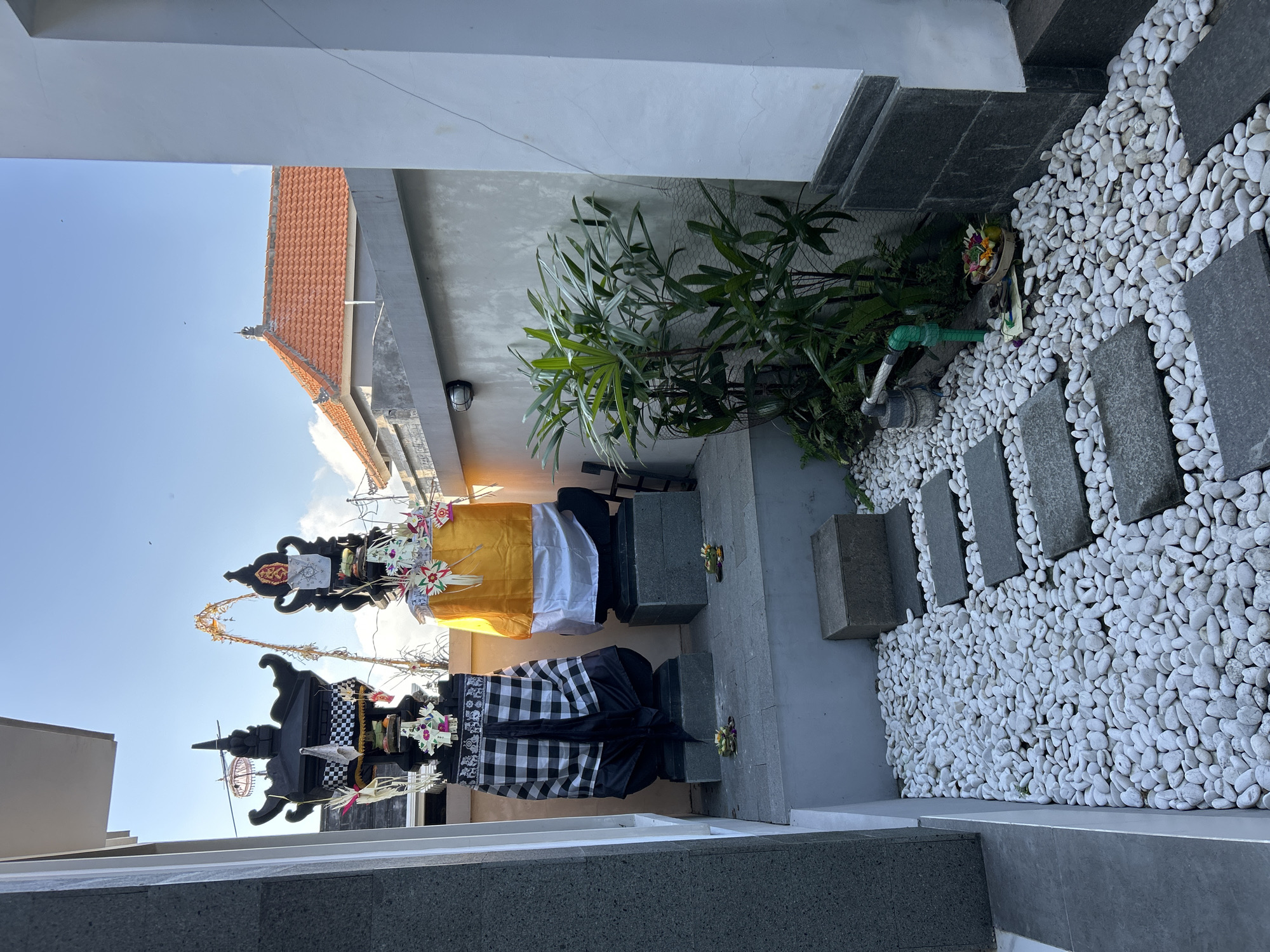 The image depicts a scene in Sukawati, Indonesia, likely a courtyard or an outdoor area adjacent to a building. Two elaborate, dark, tiered structures that appear to be ceremonial offerings or effigies are the central focus. They are draped with fabric, including a prominent yellow cloth and a black and white checkered pattern. Both structures are adorned with decorative elements, such as paper fans and floral arrangements.
The setting features a light-colored wall and a portion of a red-tiled roof visible against a bright blue sky with scattered clouds. To the right, a pathway is created with flat gray stones interspersed within a bed of smooth, white pebbles. A lush green plant is growing in this area, alongside some gardening equipment like a green pipe and a water spigot. The overall ambiance suggests a preparation for or the aftermath of a cultural or religious event. The lighting indicates it is daytime, likely morning or afternoon, with clear weather. No people are visible in the frame. The style of the offerings and the setting are consistent with Balinese traditions.