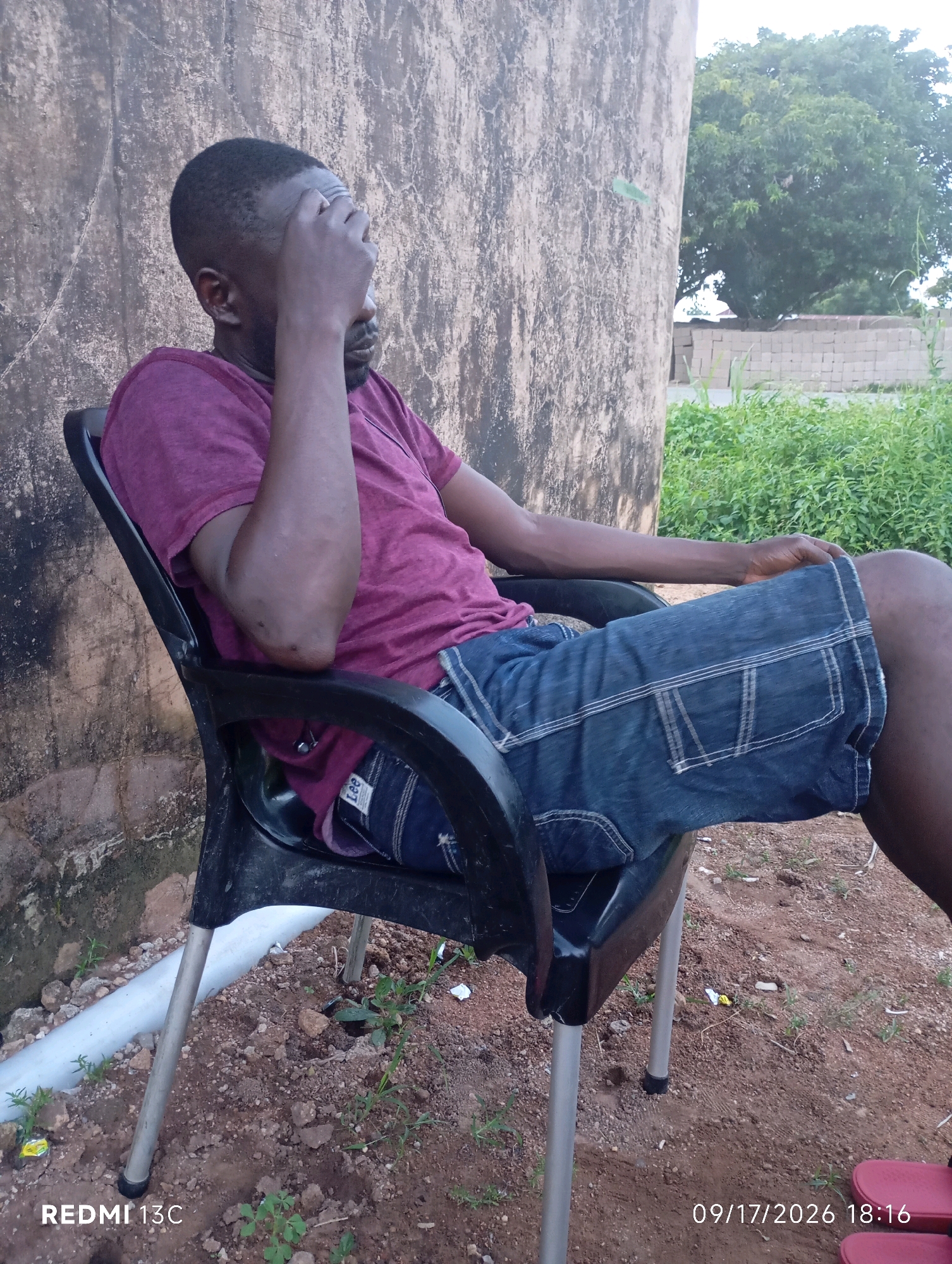 A man wearing a maroon t-shirt and denim shorts sits in a black plastic chair outdoors. He is seen from the side, with his right hand covering his forehead. The background consists of a textured, light-colored wall, green vegetation, and a pile of bricks. The ground is dirt and gravel. The image contains a timestamp and the text "REDMI 13C".