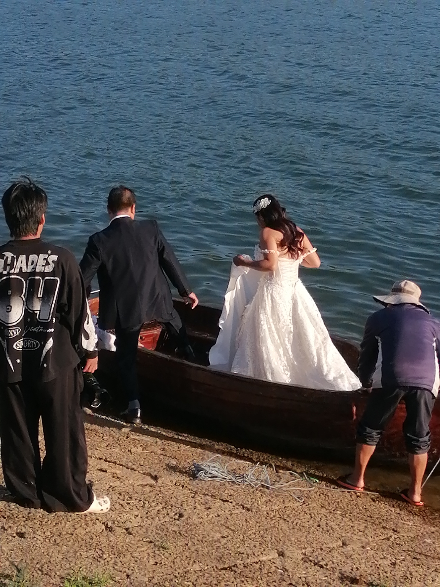 In Da Lat, Vietnam, a bride in a white lace wedding dress stands in a wooden boat on a body of water. She is being assisted by a man in a dark suit. Another man in black pants and a black shirt with graphics is standing on the shore to the left, holding a camera. A third man in a blue jacket and a hat is in the boat on the right. The water appears calm and blue, and the shore is covered in small stones and pebbles. The scene suggests a wedding photo shoot or a bridal procession on the water.
