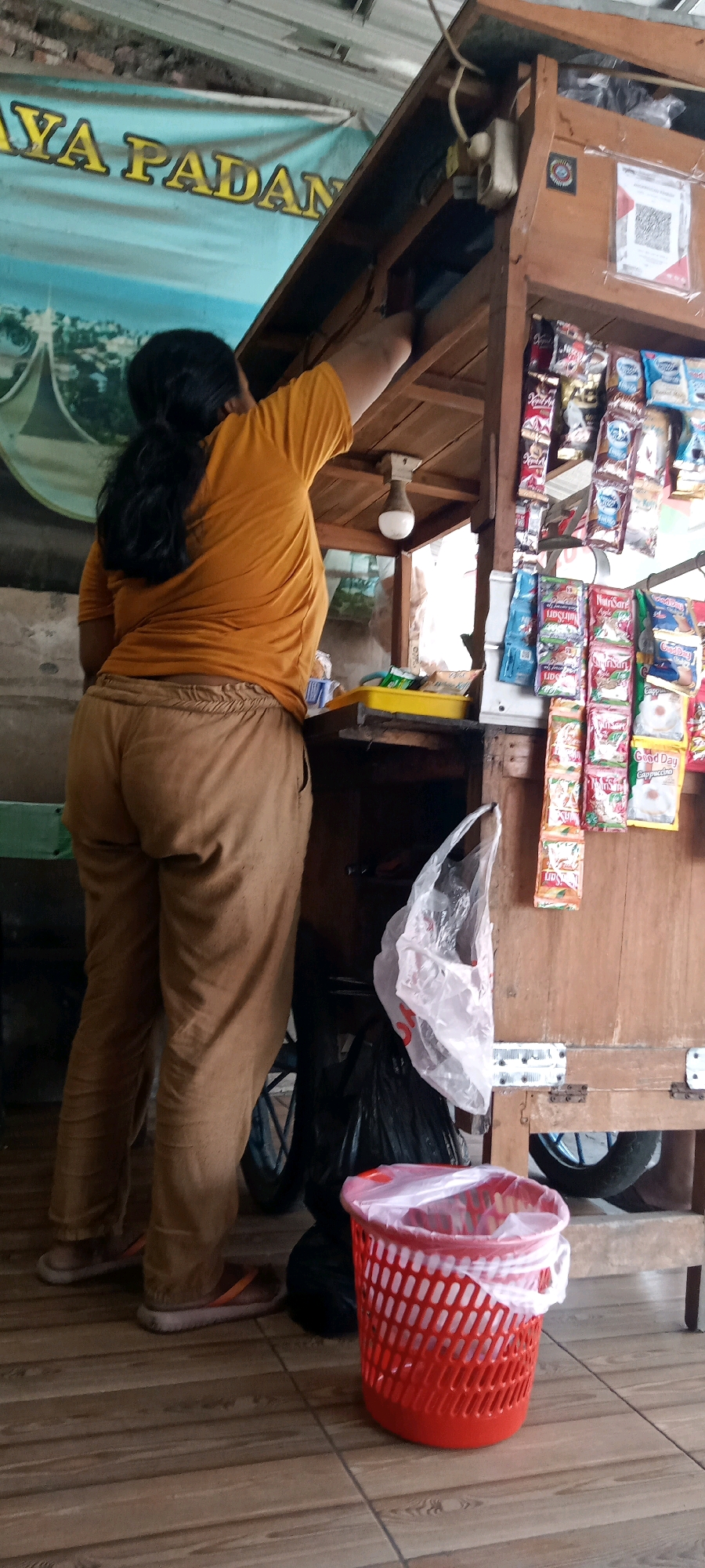 A woman with dark hair pulled back in a ponytail is standing next to a wooden stall, which appears to be a small convenience store or roadside vendor. She is wearing an orange t-shirt and brown pants. Her right arm is raised, reaching into the stall. The stall is stocked with various packaged goods, primarily food and beverage items, displayed on shelves. In front of the stall, on the tiled floor, is a red plastic trash can lined with a white plastic bag. Next to the trash can is a black trash bag. A bicycle wheel is partially visible behind the stall. In the background, a banner with yellow lettering is displayed. The scene is likely located in Delanggu, Indonesia.