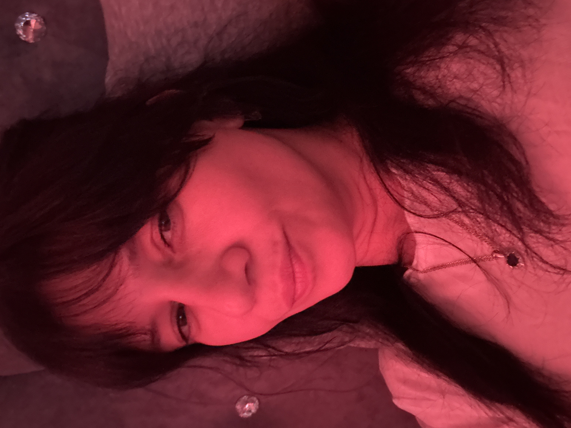 A person with long, dark hair is lying down. The lighting is red, giving a red hue to the skin and everything in the scene. The person is wearing a white shirt or top and a necklace with a pendant. The person is lying on a textured surface that looks like it may be a couch or bed. There is no other information that can be ascertained from the image.