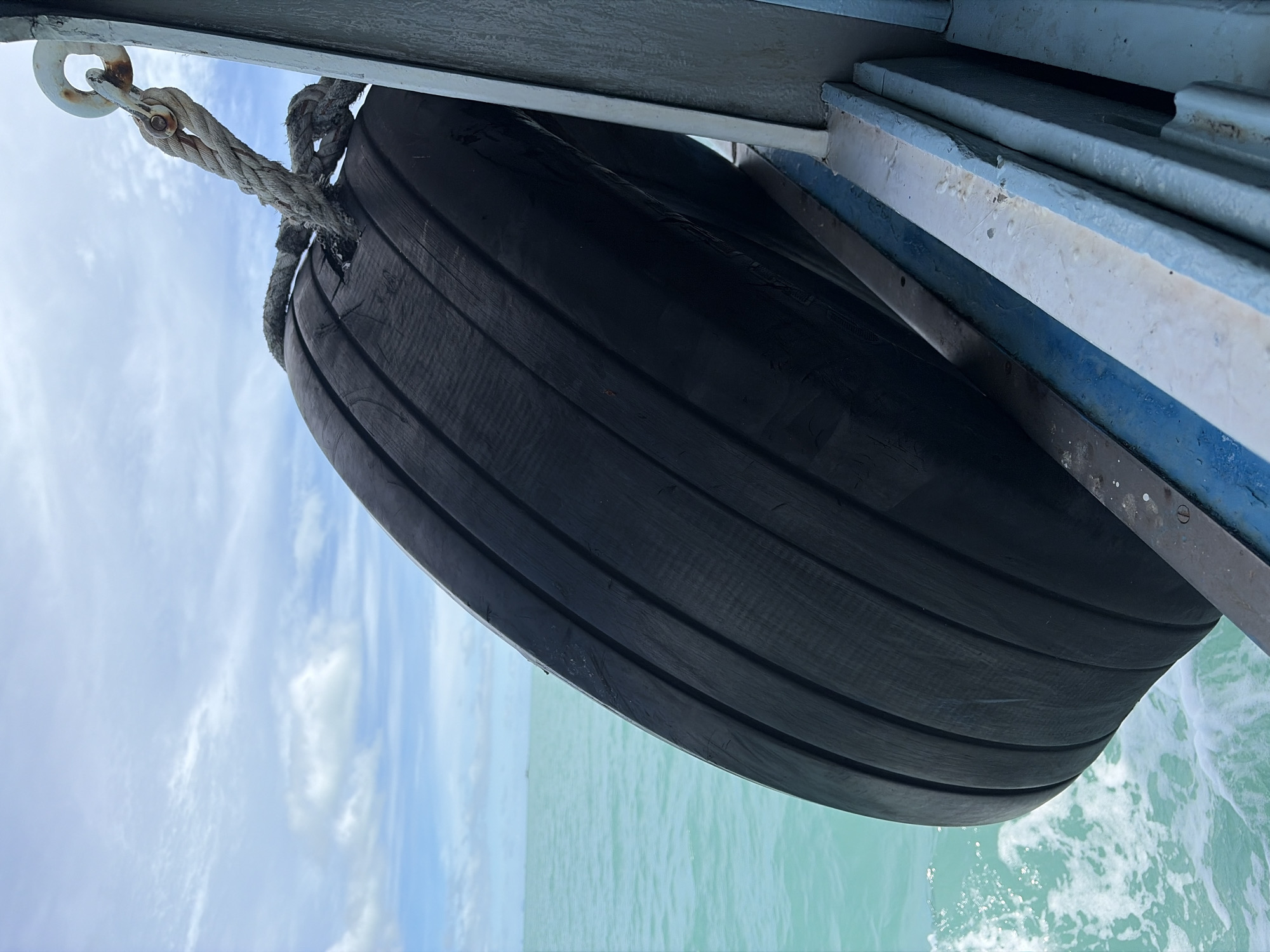 The image captures a close-up view of a large, black, cylindrical fender attached to the side of a boat or pier. The fender is made of rubber with horizontal ridges, suggesting it is designed to cushion impacts. It is secured by a thick, weathered rope and a metal fitting to an overhead structure that appears to be part of a boat's railing or a pier.
The background reveals a bright, cloudy sky and a calm, turquoise body of water, hinting at a tropical or coastal location. The water exhibits a gentle ripple, with some white foam visible near the bottom right, indicating movement. The overall lighting suggests it is daytime, likely late morning or afternoon, with the sun casting a soft glow.
The scene appears serene and practical, focused on the protective function of the fender. There is no visible text within the image. The setting is outdoors, by the water, likely in a location where boats dock or travel, such as a harbor or a coastal route. The location context provided, Baan Bang Rak, Thailand, reinforces the idea of a tropical maritime environment.