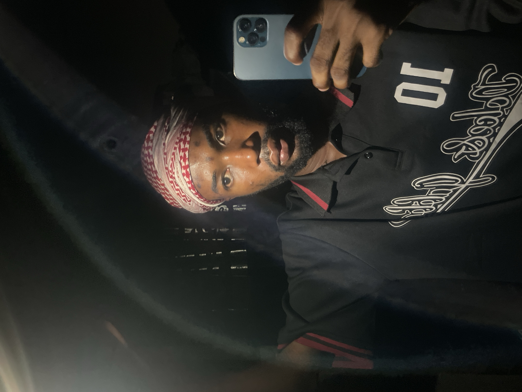 A man takes a selfie in a dimly lit environment. He wears a red and white patterned headscarf and a black collared shirt with red stripes on the shoulders and the number "10" on the chest. He holds a light blue smartphone in his right hand, which is positioned above his head. The environment appears to be dark and possibly enclosed, with some reflections indicating a smooth surface. The location is unknown and not identifiable.