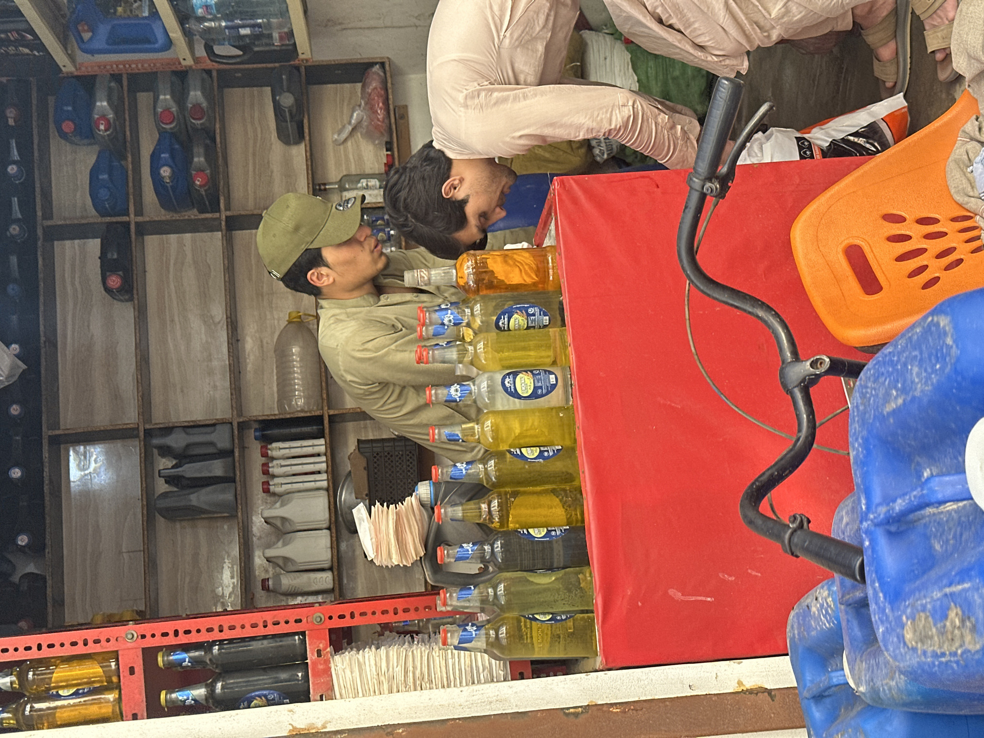 This image captures a scene within what appears to be a small shop or stall, potentially selling lubricants or other bottled liquids, with the image itself oriented ninety degrees clockwise. Two men are featured. One man, wearing a green cap and a light-colored long-sleeved shirt, stands on the left, looking right and slightly downwards towards items on a red counter. The second man, dressed in a light-colored long-sleeved garment, is positioned on the right side of the counter, bending over with his head down, also looking at the products.
The activity suggests an interaction or inspection of goods. Numerous clear glass and plastic bottles, filled with liquids of varying colors (yellowish, clear, dark), are neatly arranged on the red counter. Some bottles feature blue and white labels, with "BINA" and "AL-ASRA" being clearly visible brand names. Behind these bottles, stacks of white folded papers, possibly napkins or small bags, are visible.
The shop interior features shelves made of both wood and metal. On the wooden shelves in the background, various plastic containers, some blue, others grey or black, are stored. Some grey containers on a shelf have "000" printed on them. On the far right, a black bicycle handlebar with a brake lever is visible, positioned near the red counter and next to an orange plastic object, possibly a child's seat or a small utility item. Several large blue plastic jerricans are partially seen in the lower right corner. The lighting is even, suggesting an indoor setting or a covered outdoor stall. The men's expressions appear neutral and focused on the products.