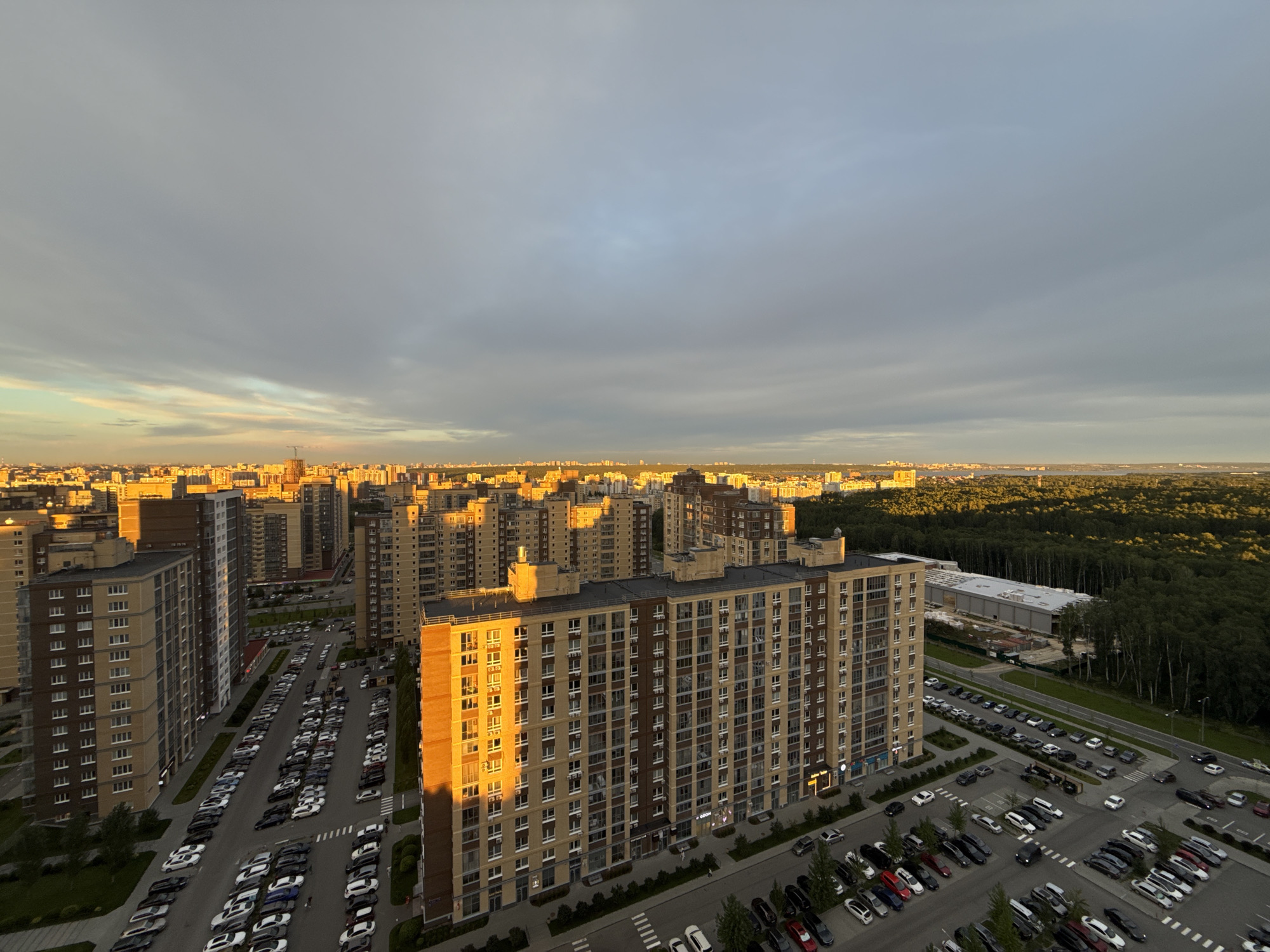 The image captures an aerial view of a modern urban landscape at sunset. The foreground is dominated by several multi-story residential buildings with balconies and numerous windows. A large parking lot filled with a variety of cars is visible between the buildings. The cars are parked in rows, with a mix of colors and models. Beyond the immediate residential area, the city extends into the distance, characterized by more buildings and a dense forest. The sky is partly cloudy, with the warm hues of sunset casting a golden glow on the buildings and illuminating the clouds. The lighting suggests it is late afternoon or early evening. The overall impression is of a developing urban area bordered by a natural wooded expanse, bathed in the soft light of sunset. The location is a residential district in Chelyabinsk, Russia, likely a newly developed area on the outskirts of the city. There are no visible people in the scene, and the focus is on the architecture, the parked vehicles, and the environmental setting.