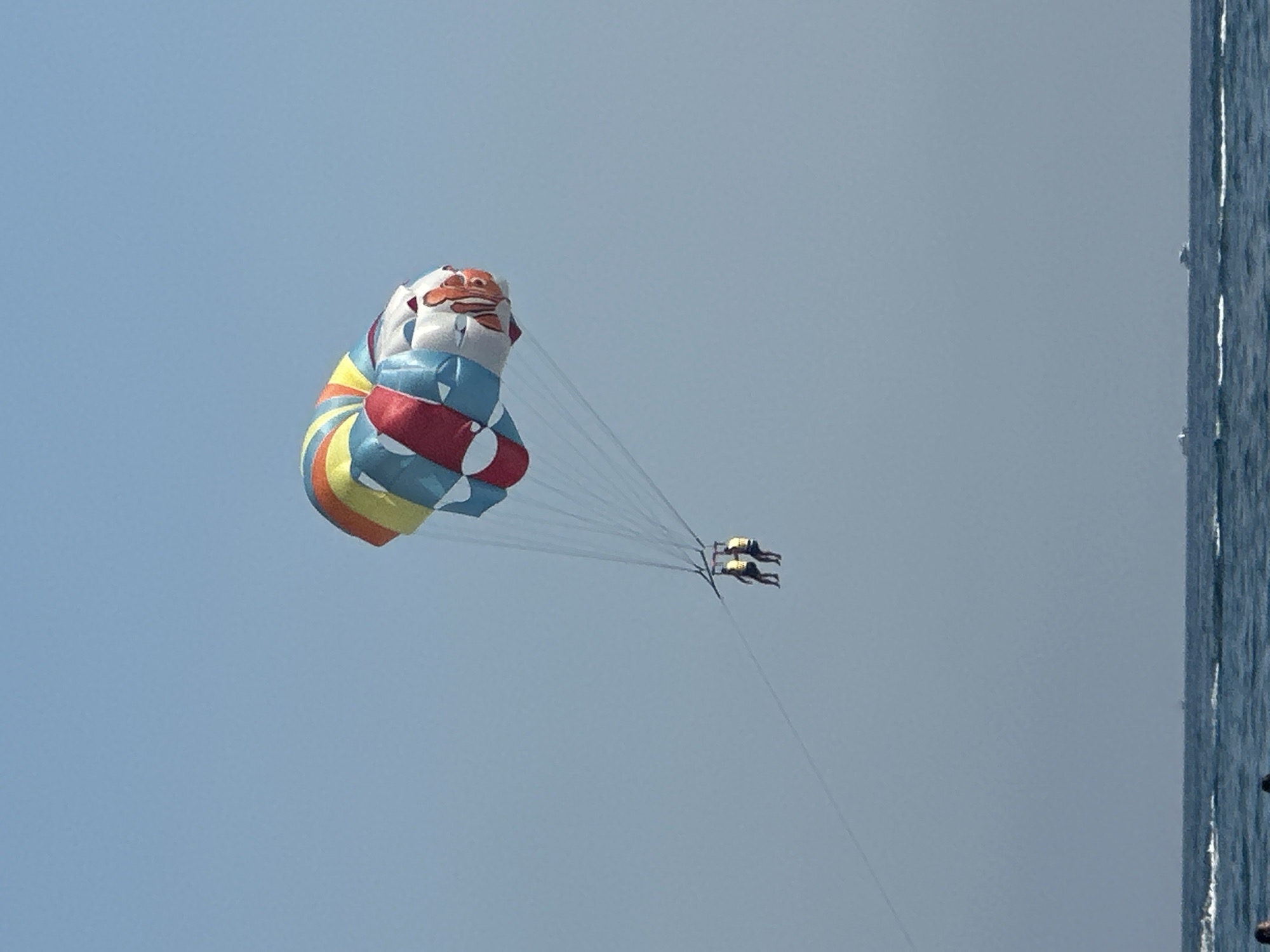 Two individuals are parasailing in the sky above the Adriatic Sea. The colorful parachute, featuring segments of red, white, blue, yellow, and orange, with a clownfish graphic, is high in the clear blue sky. The two people are harnessed and suspended beneath the parachute, appearing to be enjoying the activity. Below them, the calm blue sea stretches to the horizon, with several boats visible in the distance. The shoreline is not visible in this image, suggesting the activity is taking place offshore from the city of Boreti, Montenegro.