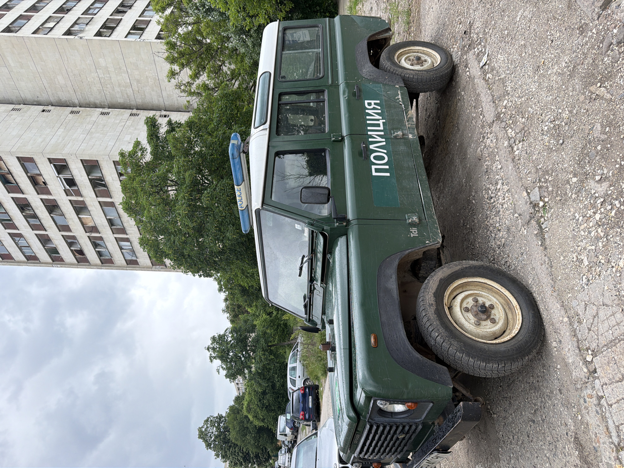 The image shows a green Land Rover Defender police vehicle, likely of Bulgarian origin, parked on a gravel and concrete surface next to a street. The vehicle has a white roof and the word "ПОЛИЦИЯ" (Police) is printed in white Cyrillic letters on its side. A blue light bar is mounted on the roof. Several other cars are parked along the street behind the Land Rover. In the background, a tall, concrete building with many windows can be seen, along with some trees. The weather appears overcast. While Pleven can be observed from the license plate on the car, the precise street location is not evident from the image.