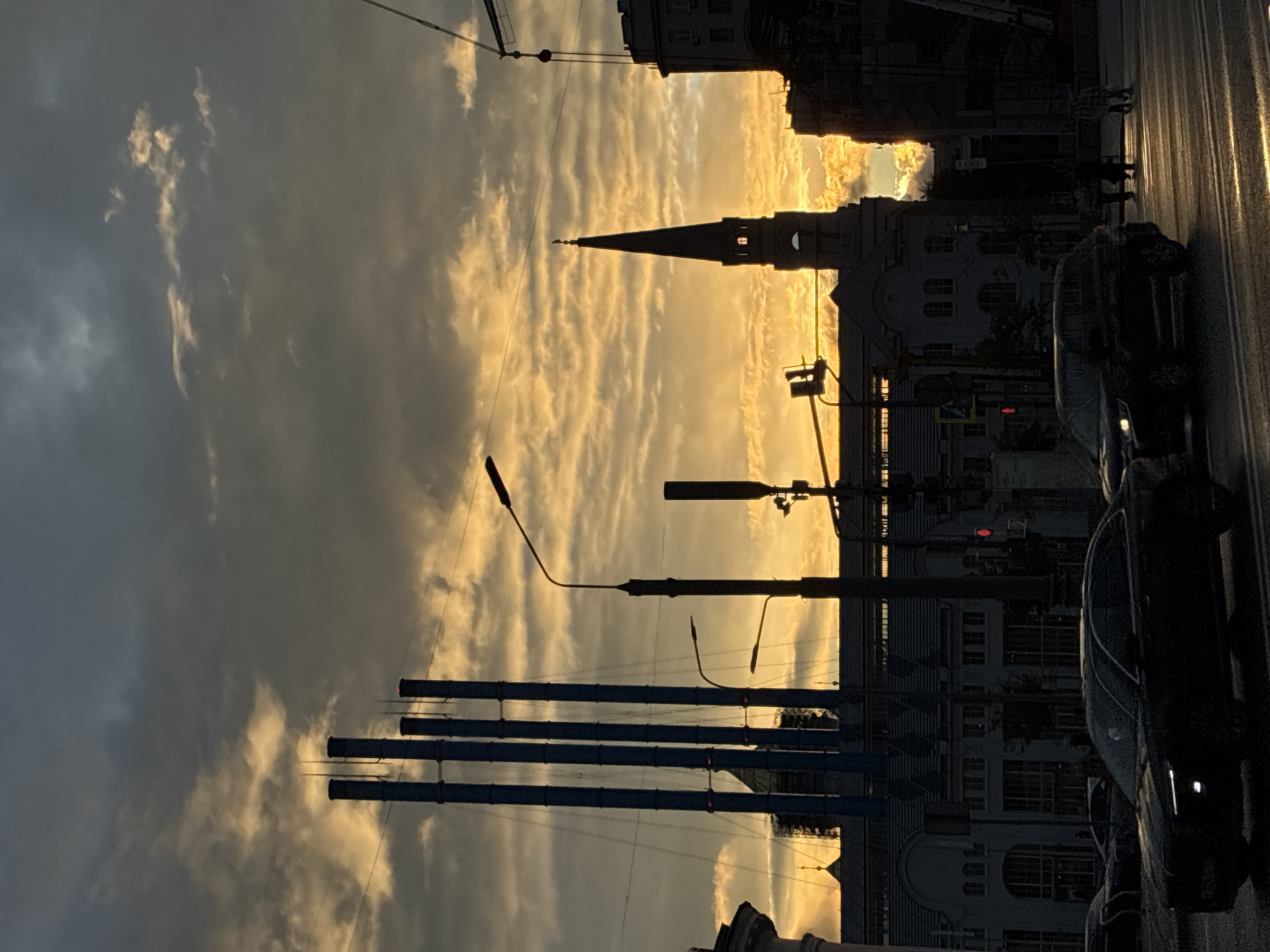 The image shows a street in Moscow, Russia, at sunset. A tall church spire is silhouetted against the golden sky. To the left, there are tall, cylindrical industrial structures. Streetlights and traffic signals are visible. Cars are parked along the right side of the street, and a couple of people are walking on the sidewalk. The sky is filled with clouds that are brightly lit by the setting sun. The overall scene is a blend of urban and industrial elements under a dramatic sky.