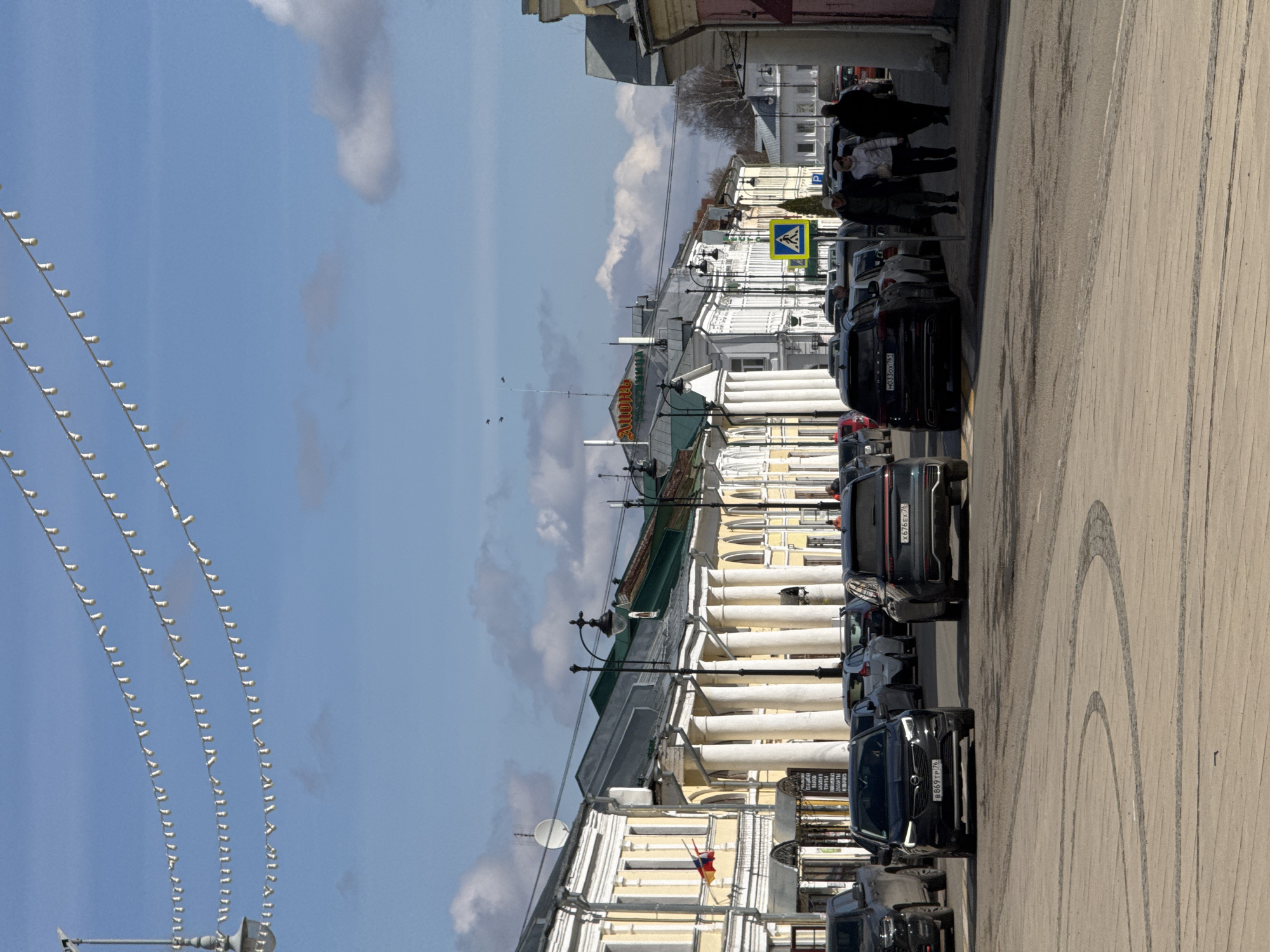 The image depicts a street in Novo-Nikol'skoe, Russia, lined with classic buildings featuring white columns. Cars are parked along the right side of the street, with several dark-colored SUVs visible. People are walking on the sidewalk on the right side of the image. Overhead, strings of decorative lights are strung across the street. The sky is blue with scattered clouds. A crosswalk sign is visible. There are tire marks on the road.