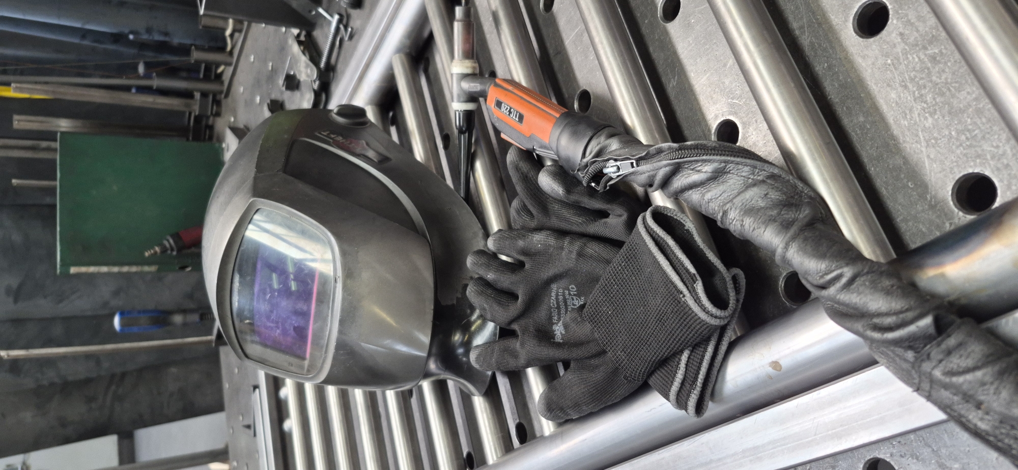 This is a close-up, slightly angled, eye-level shot of welding equipment on a metal workbench. In the foreground and center, a dark gray welding helmet rests on a pile of silver metal tubes. To the left of the helmet, a pair of black fabric gloves is stacked. Extending from beneath the helmet and gloves is the orange and black handle of a welding torch, wrapped in black leather shielding. The background shows a blurred industrial environment with metal structures and a green cabinet. The workbench surface is a grid of metal tubes with circular holes. The scene appears to be in a workshop in Czaple, Poland.
