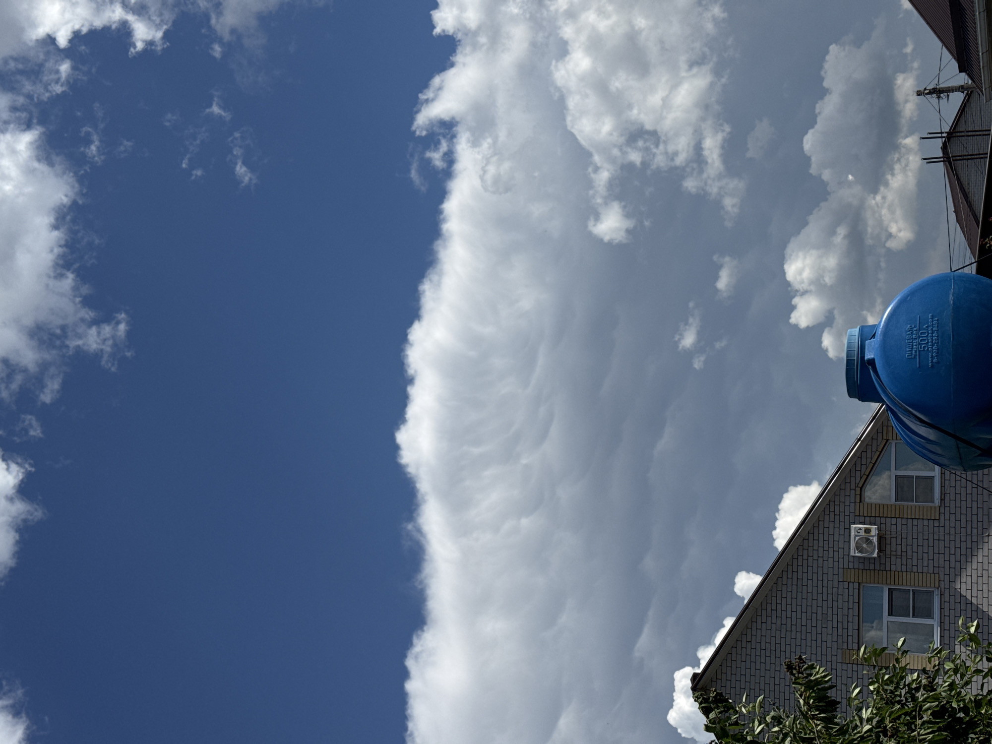 This image shows a low-angle view of the sky above a building and some foliage. The sky is a deep blue with dramatic white clouds. One large cloud formation stretches across the frame, appearing textured and somewhat turbulent, with a distinct line separating it from the clearer blue sky on the left. In the lower right corner, the top portion of a grey brick building is visible. It has a pitched roof and two windows with light-colored frames. An air conditioning unit is mounted on the wall between the windows. To the right of the building, a large, round, blue plastic tank is partially visible. Text on the blue tank reads "ПИЩЕВАЯ 500л "Plast Tank" www.plast-tank.ru +7(800) 250-93-23". The lighting suggests it is daytime, likely mid-morning or afternoon, with bright sunlight casting shadows and illuminating the clouds. The overall impression is one of a bright, possibly warm day with dynamic cloud formations. The location cues suggest Mikhaylovsk, Russia. No people are visible in the image.