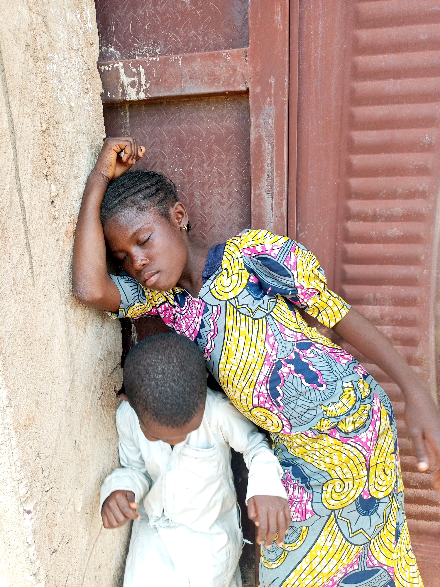 A young African girl with braided hair and closed eyes leans her head on her left arm, which is propped against a beige, textured wall. She is wearing a colorful dress with a pattern of yellow, pink, and blue swirls and geometric shapes. A young African boy, seen from behind, stands in front of the girl, his head bowed. He is wearing a light-colored tunic. They are positioned in a narrow passage between the wall and a corrugated, reddish-brown metal door. The scene appears to be in an outdoor setting, possibly a doorway or alley, within the city of Tempure, Nigeria.