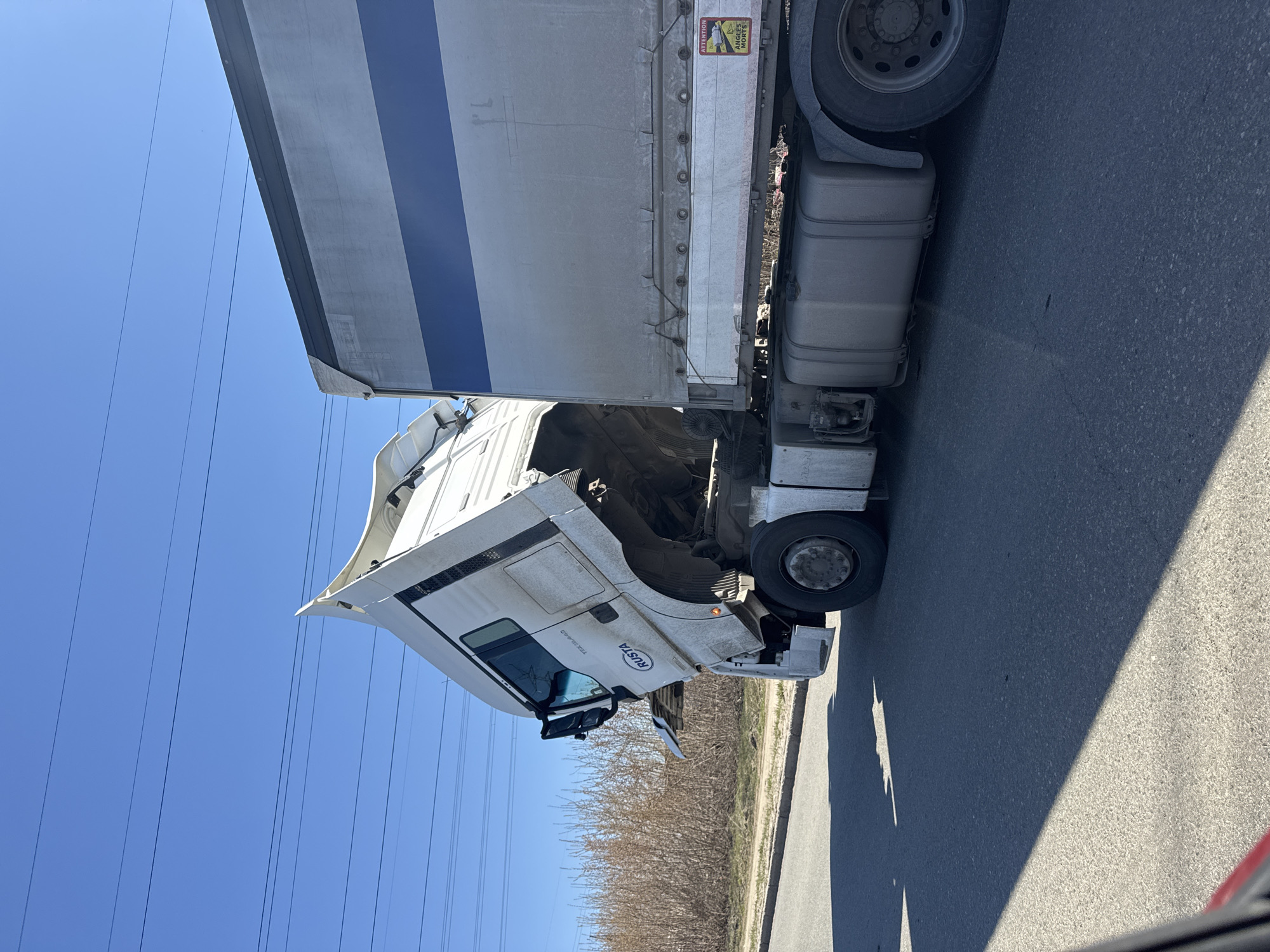The image shows a white Rusta truck with a blue striped trailer parked on an asphalt road. The truck's cab appears to be tilted forward. Overhead electrical wires are visible against a clear blue sky. A line of leafless bushes borders the road. Based on the "Rusta" branding on the truck and the provided location, the image is presumed to be in the city of Chaikovsky, Russia. There are no people visible in the image.