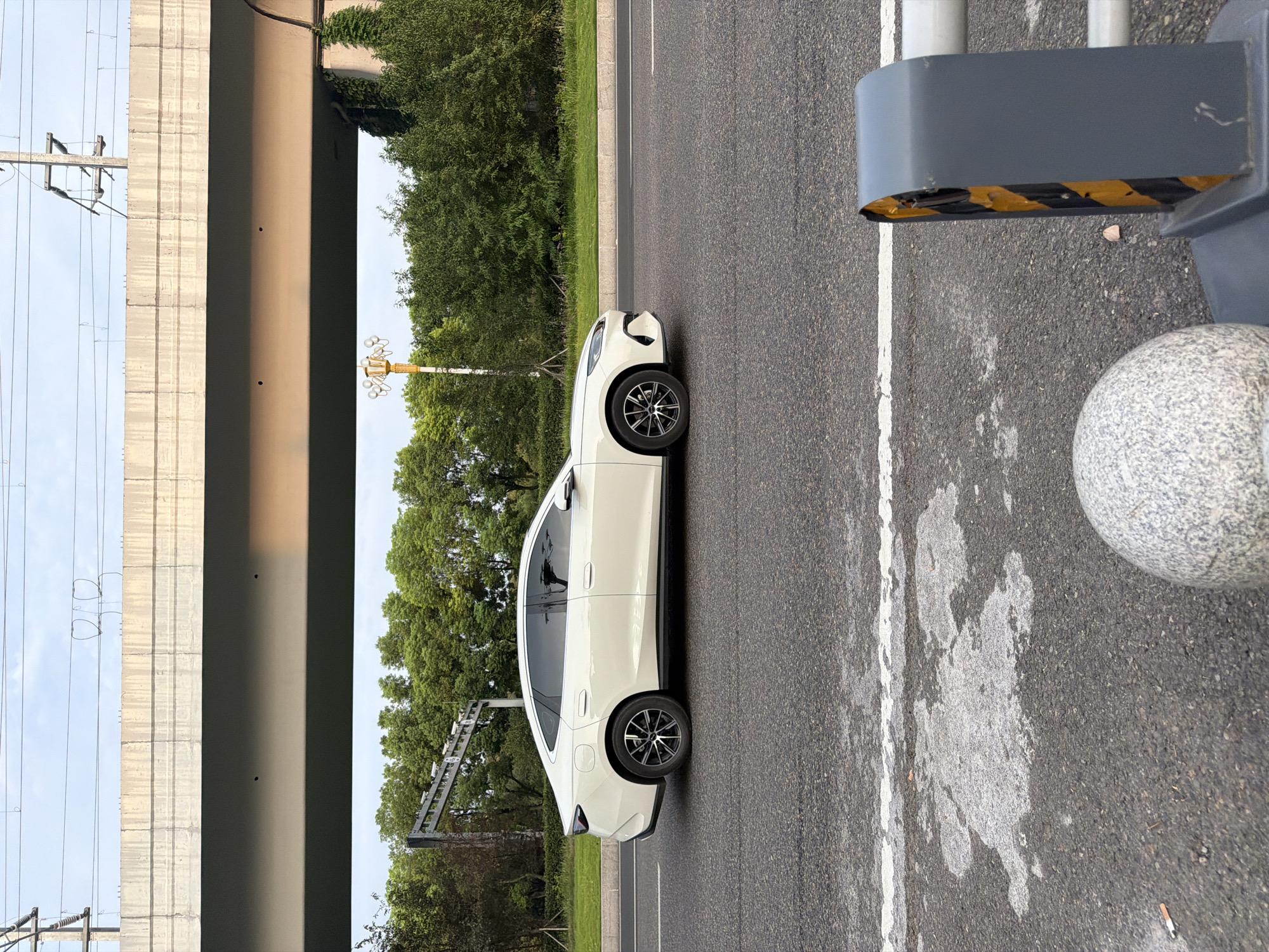 A white sedan is captured in profile, driving on an asphalt road under an overpass. The car has tinted windows and black and silver alloy wheels. In the foreground, the road surface shows wear and tear with white lane markings. A grey traffic barrier with yellow and black diagonal stripes is visible on the right side of the frame, alongside a grey spherical object. Behind the road, a row of lush green trees forms a backdrop. Above the trees, a concrete overpass spans the scene, with power lines and poles visible against a pale blue sky. The scene is indicative of a road in the city of Jiangjin, China, possibly during daytime.