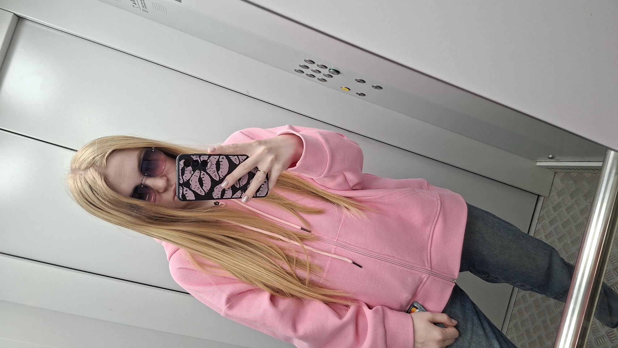 A young woman with long blonde hair and wearing sunglasses is taking a selfie in what appears to be an elevator. She's wearing a pink hoodie and jeans. The phone case has a pattern of pink lips on a black background. Her right hand is holding the phone, and she wears a gold ring on her ring finger. The elevator has white walls, a stainless steel handrail, and a patterned metal floor. Elevator buttons are visible above her. The location is Chaikovsky, Russia.