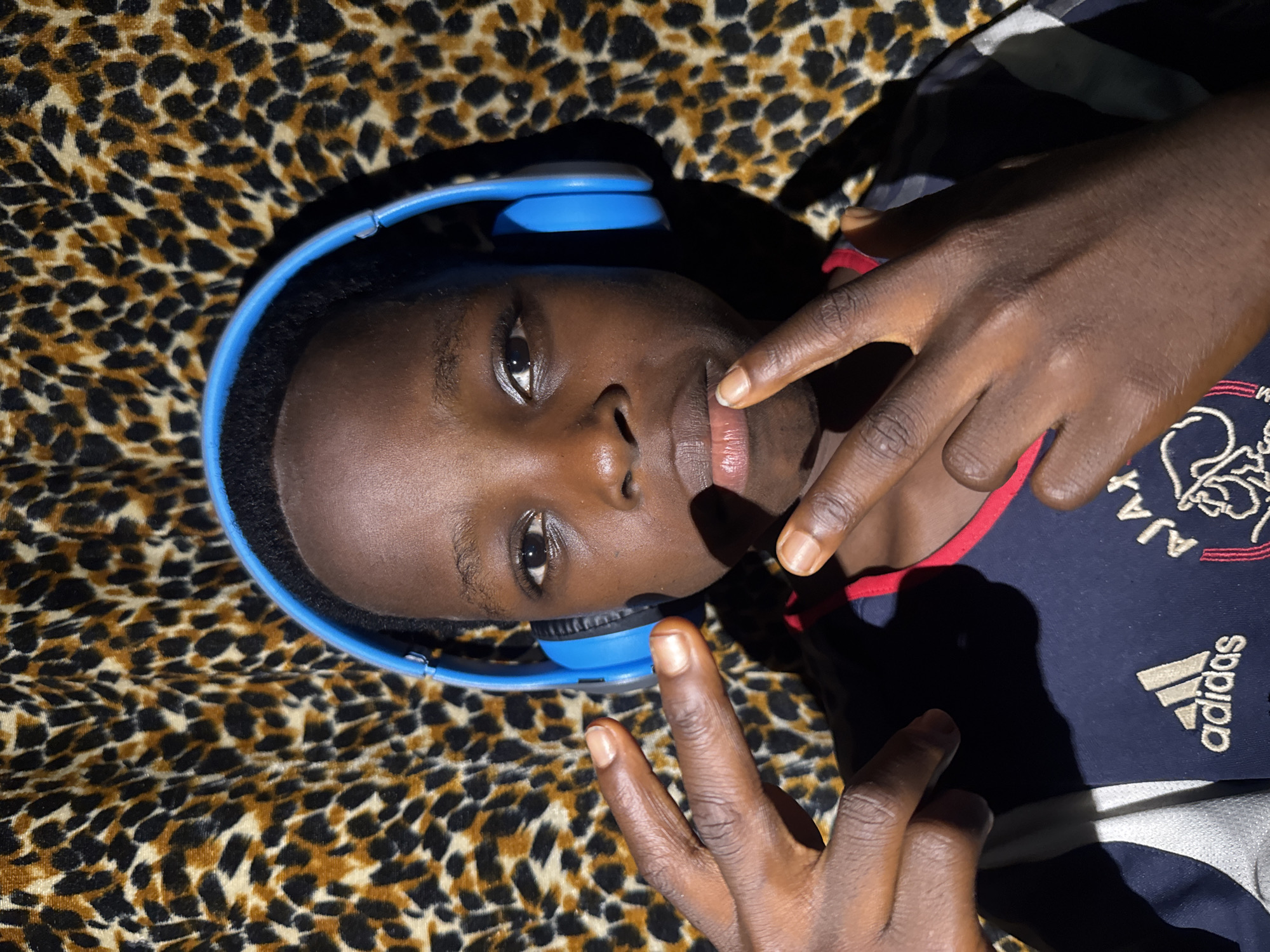 A young person of African descent is lying down on a leopard-print surface, presumably fabric, wearing blue headphones. The person is facing the camera. One hand is positioned near their mouth with the index finger touching the lower lip, while the other hand is positioned below the face, fingers pointed upwards. The person is wearing a dark blue Adidas shirt with some faded white detailing. There is insufficient information to place the scene in Onitsha, Nigeria.
