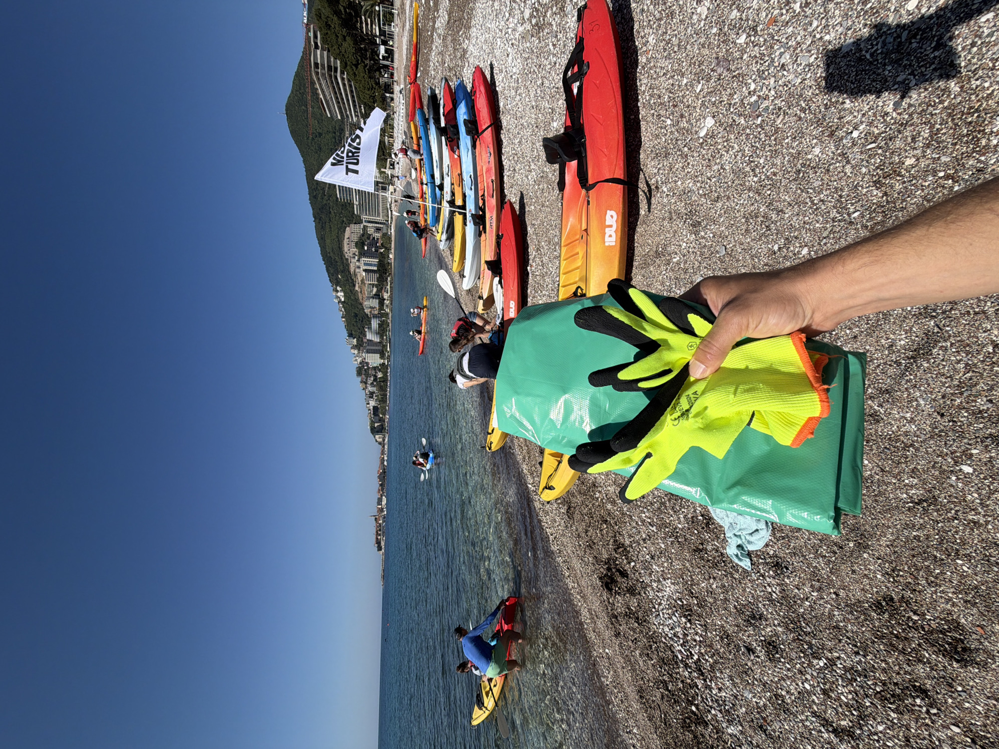 The image shows a pebble beach on a sunny day. In the background, a coastline with buildings on a steep hill is visible, likely in the city of Budva, Montenegro. Several colorful kayaks are lined up on the beach, some with people nearby or paddling in the clear blue water. In the foreground, a person's arm holds a green bag with yellow and black gloves. The overall scene suggests a kayaking tour operation.