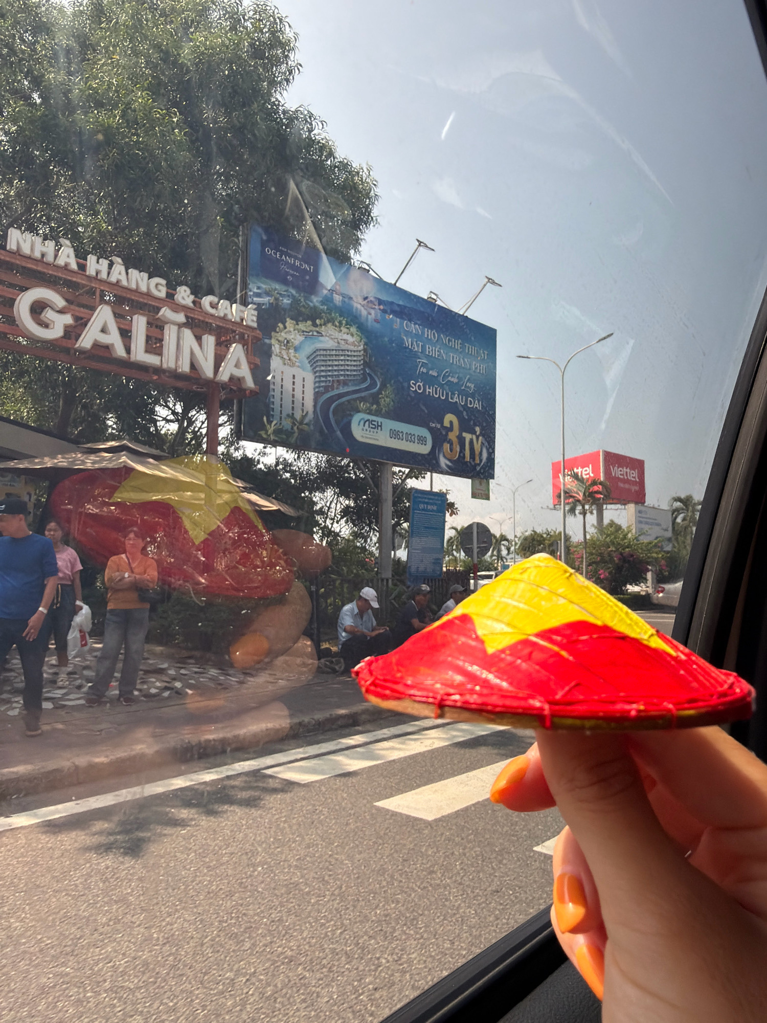 In Bắc Cam Ranh, Vietnam, a person's hand, adorned with bright orange nail polish, holds a miniature conical hat, a symbol of Vietnam, painted red and yellow with a star design, up to the camera. The hat is positioned in the foreground, obscuring a portion of the scene behind it. The background reveals a street scene viewed from inside a vehicle, indicated by the tinted window and the car's interior framing the shot. To the left, a large, white sign with the words "NHÀ HÀNG & CAFE GALINA" is visible, suggesting a restaurant or cafe. Next to it, a large billboard advertises "OCEANFRONT." Further in the background, a red billboard displays "Viettel" with its logo. Several people are casually standing or sitting along the roadside, some under a shaded awning. The environment appears to be outdoors, with trees and sunlight casting shadows. The road surface is visible with a white pedestrian crossing.
