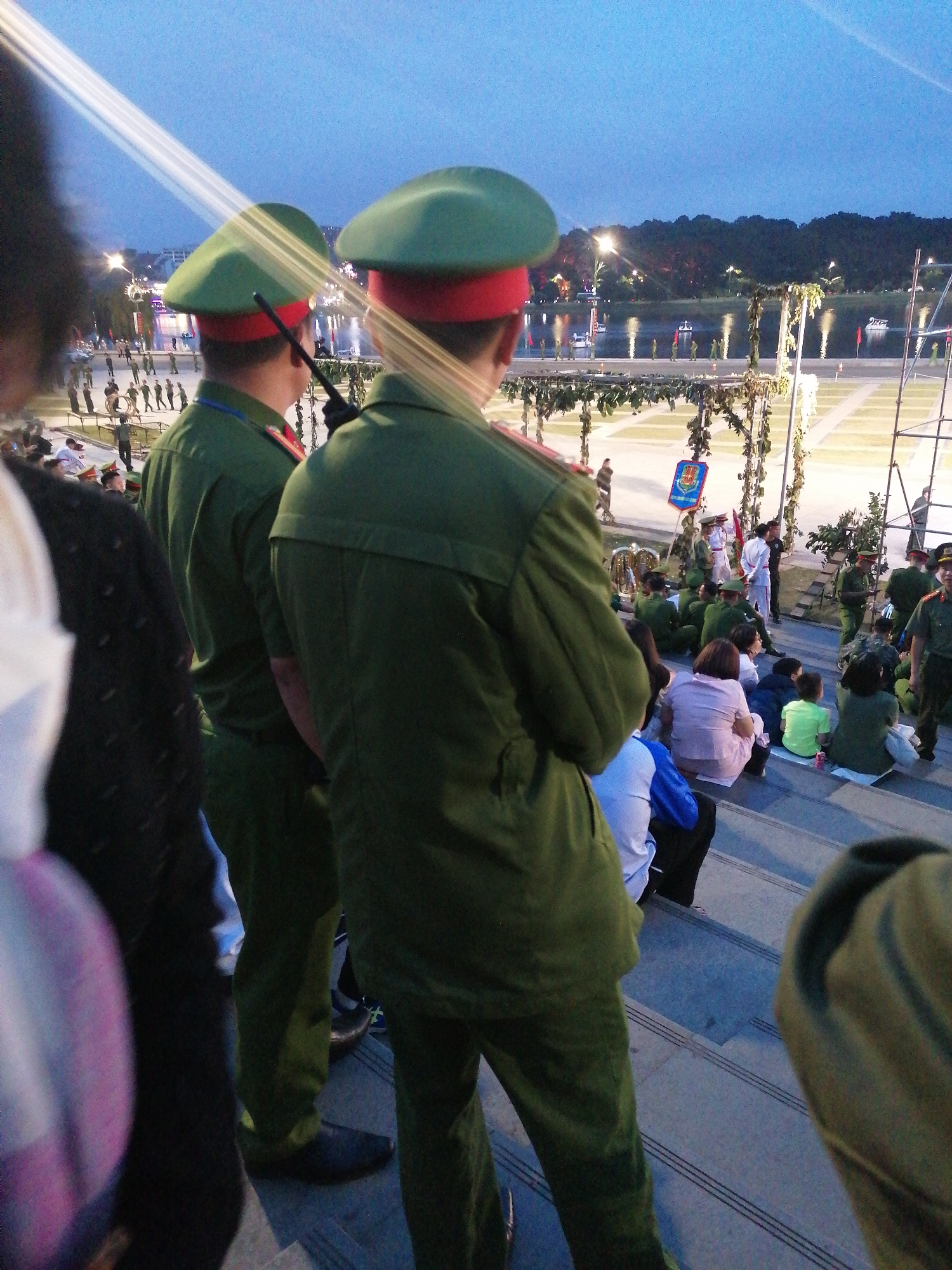 This is a medium shot of an outdoor event, likely a parade or ceremony, as indicated by the uniformed individuals and spectators. The scene is captured at dusk, with the sky a deep blue and illuminated by artificial lights. In the foreground, two men in green military-style uniforms and green berets with red bands are facing away from the camera, observing an event. They appear to be part of an honor guard or security detail. One man on the left holds a dark, slender object, possibly a rifle or baton. Behind them, a crowd of people are seated on tiered steps, also facing the direction of the observed event. They are a mix of adults and children.
Further into the background, a body of water, possibly a lake or river, is visible with several boats. The far bank of the water is lined with trees and some buildings. Lights are reflected on the water's surface, suggesting a city or resort area. A decorative archway with hanging foliage is positioned to the right of the two foreground figures. The overall atmosphere is one of organized public gathering. The presence of the lake and the general topography, combined with the style of architecture and the uniforms, suggest the location could be a public square or waterfront park in a Vietnamese city like Da Lat, Vietnam, known for its scenic lakes and French colonial architecture.